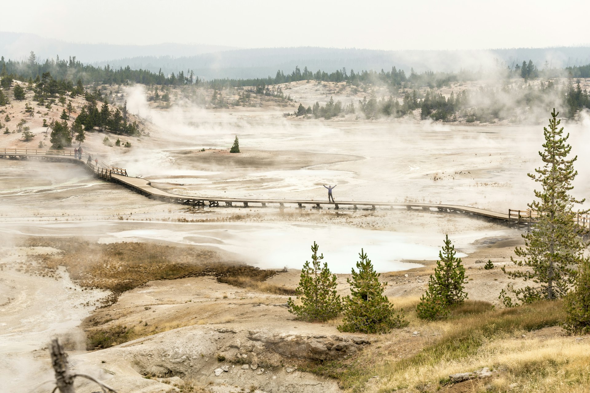 A woman holds her hands in the air and smiles as she stands alone on a boardwalk through flat landscape punctuated by geysers