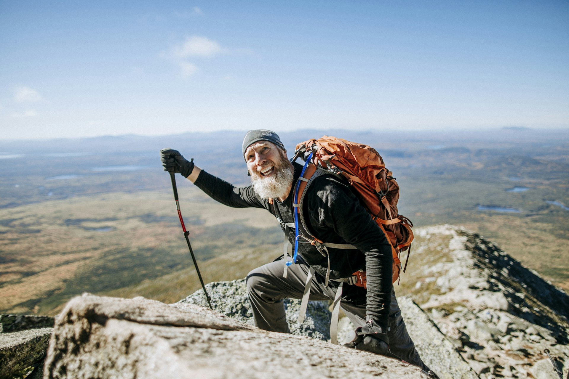 An older, bearded white man in hiking gear climbs over some rocks on a mountain in Maine, New England