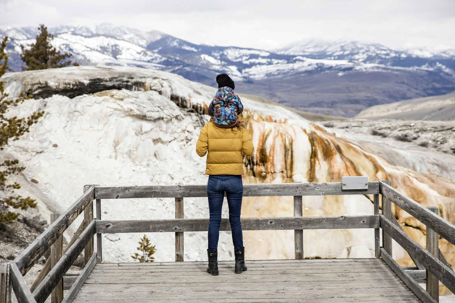 A visitor has a small child on their shoulders as they stand on a boardwalk and face out across the snowy landscape in Yellowstone National Park