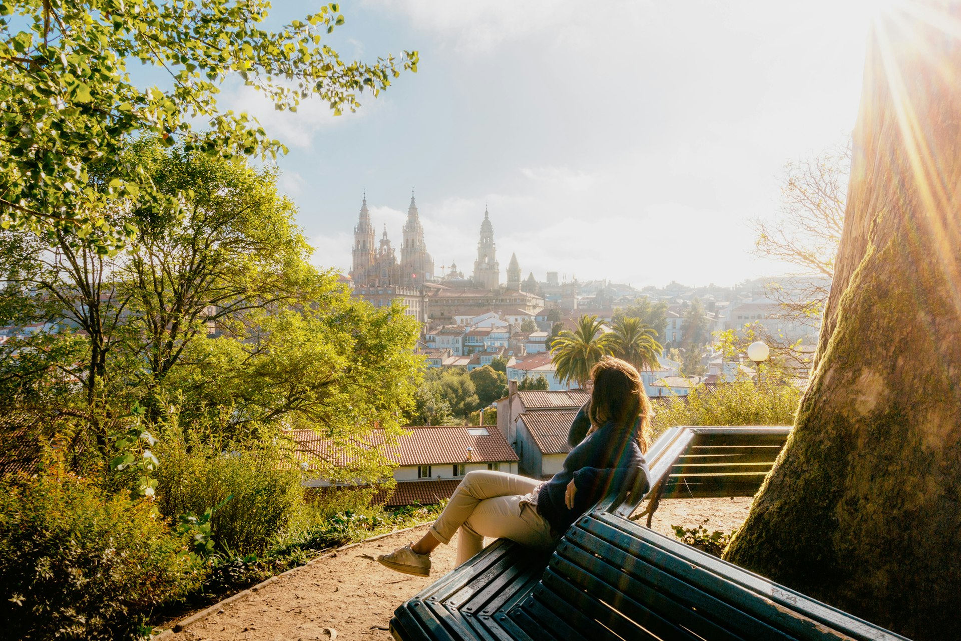 Woman on a park bench watching Santiago de Compostela Cathedral at sunrise, park and sunny sky