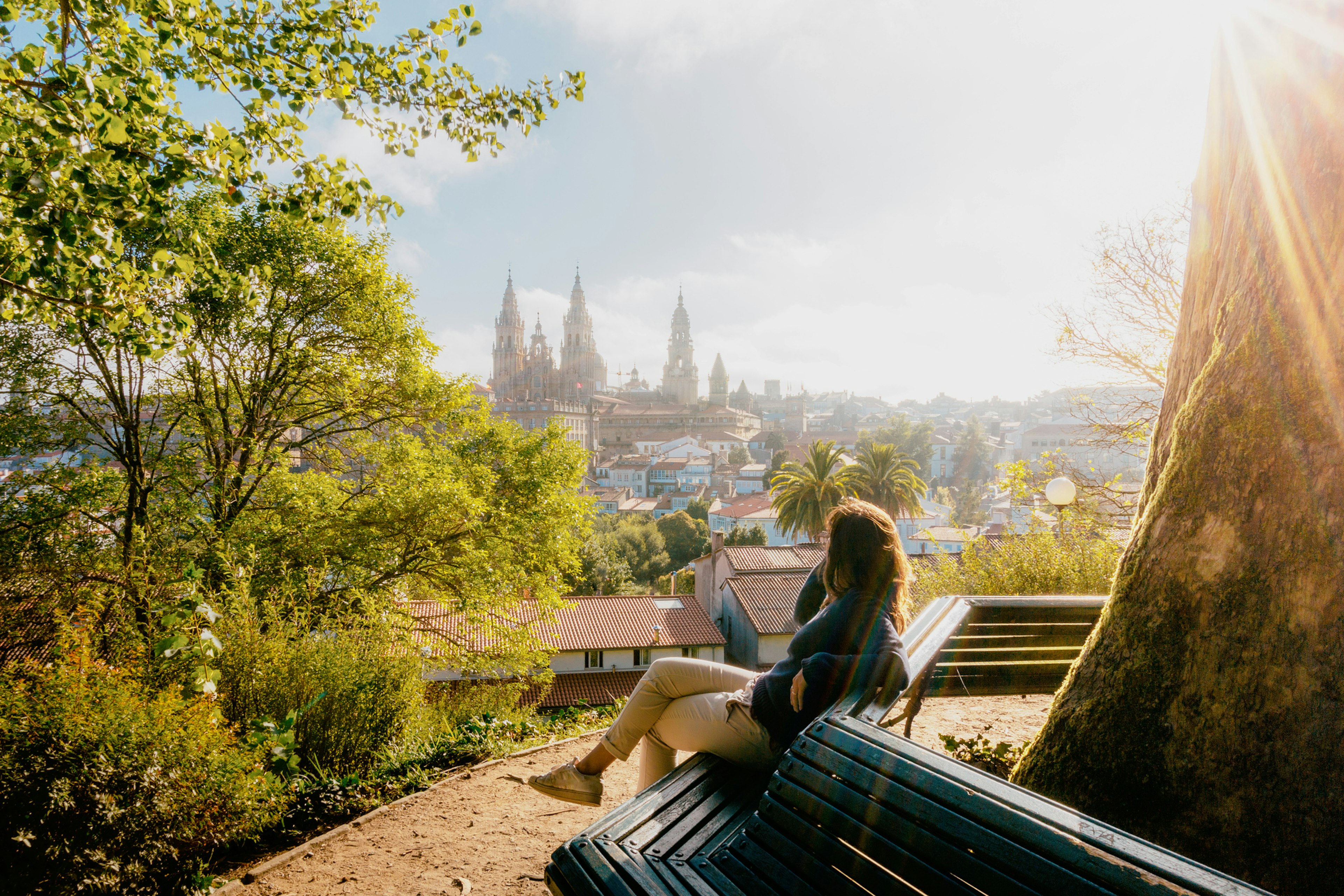 Woman on a park bench watching Santiago de Compostela Cathedral at sunrise, park and sunny sky