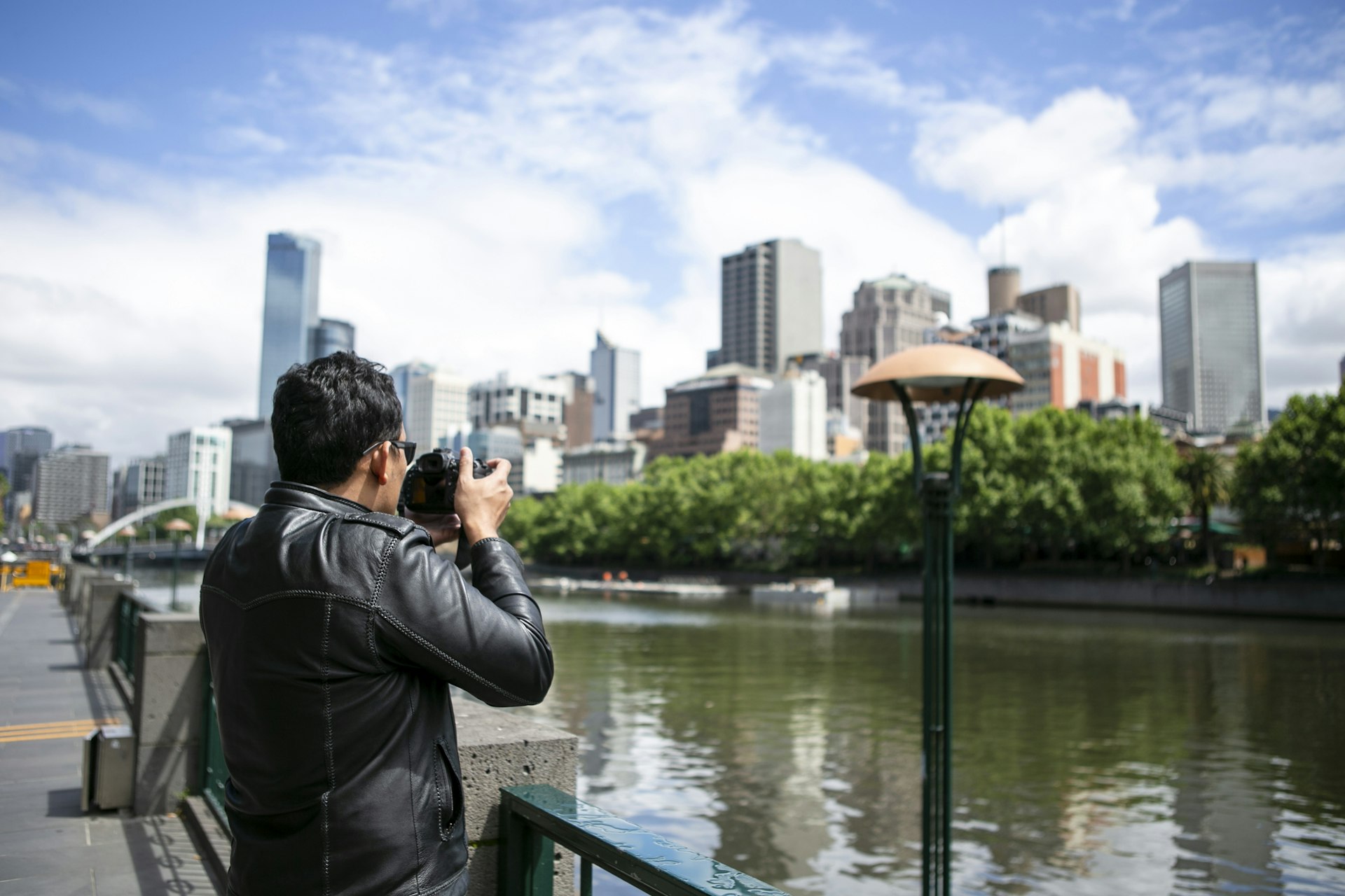 An older Asian traveler in a leather jacket stands beside of Yarra river in Melbourne city centre looking over towards the financial district taking photos