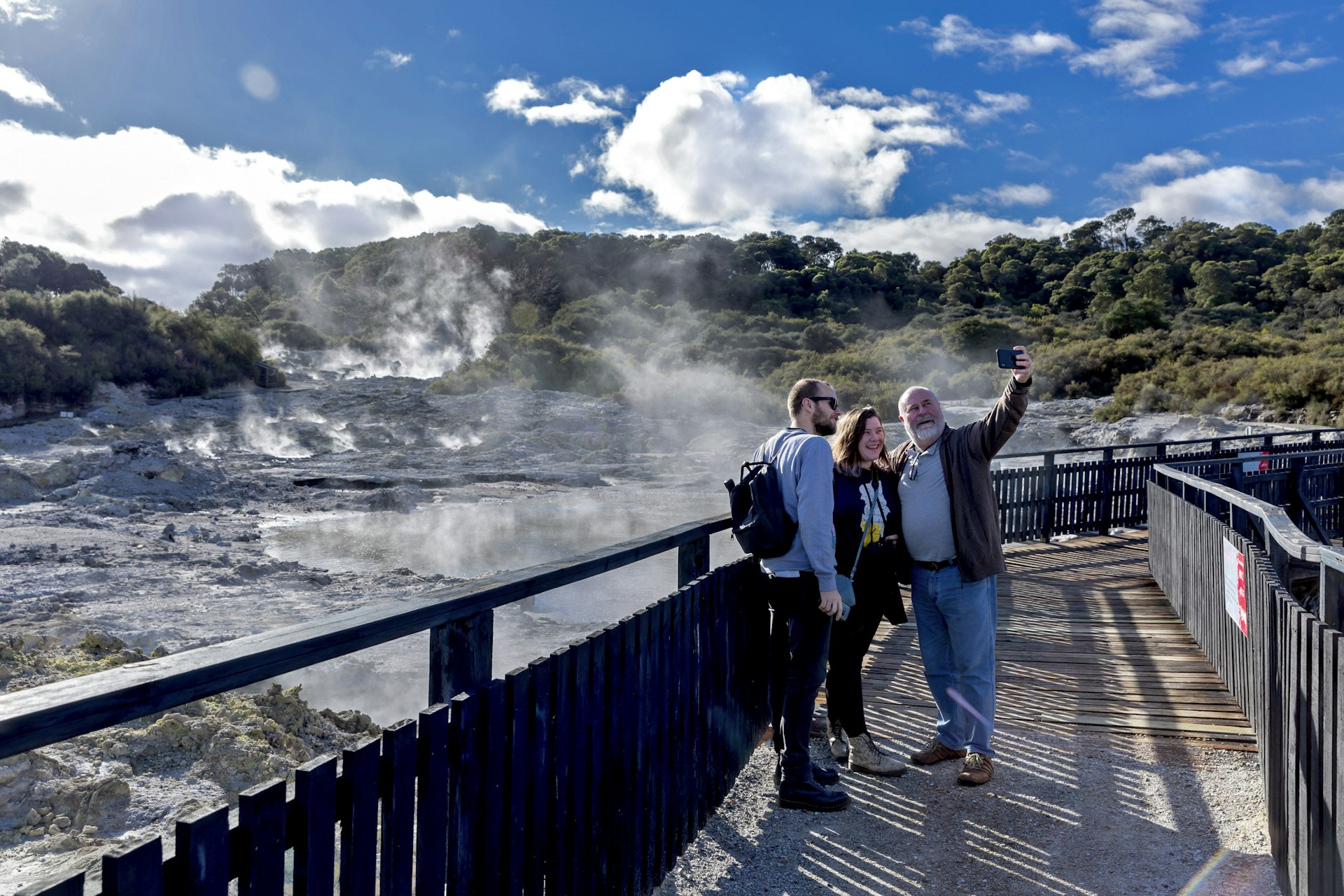Three people of differing ages stand on a boardwalk and pose for a selfie. Behind them the earth is steaming and bubbling