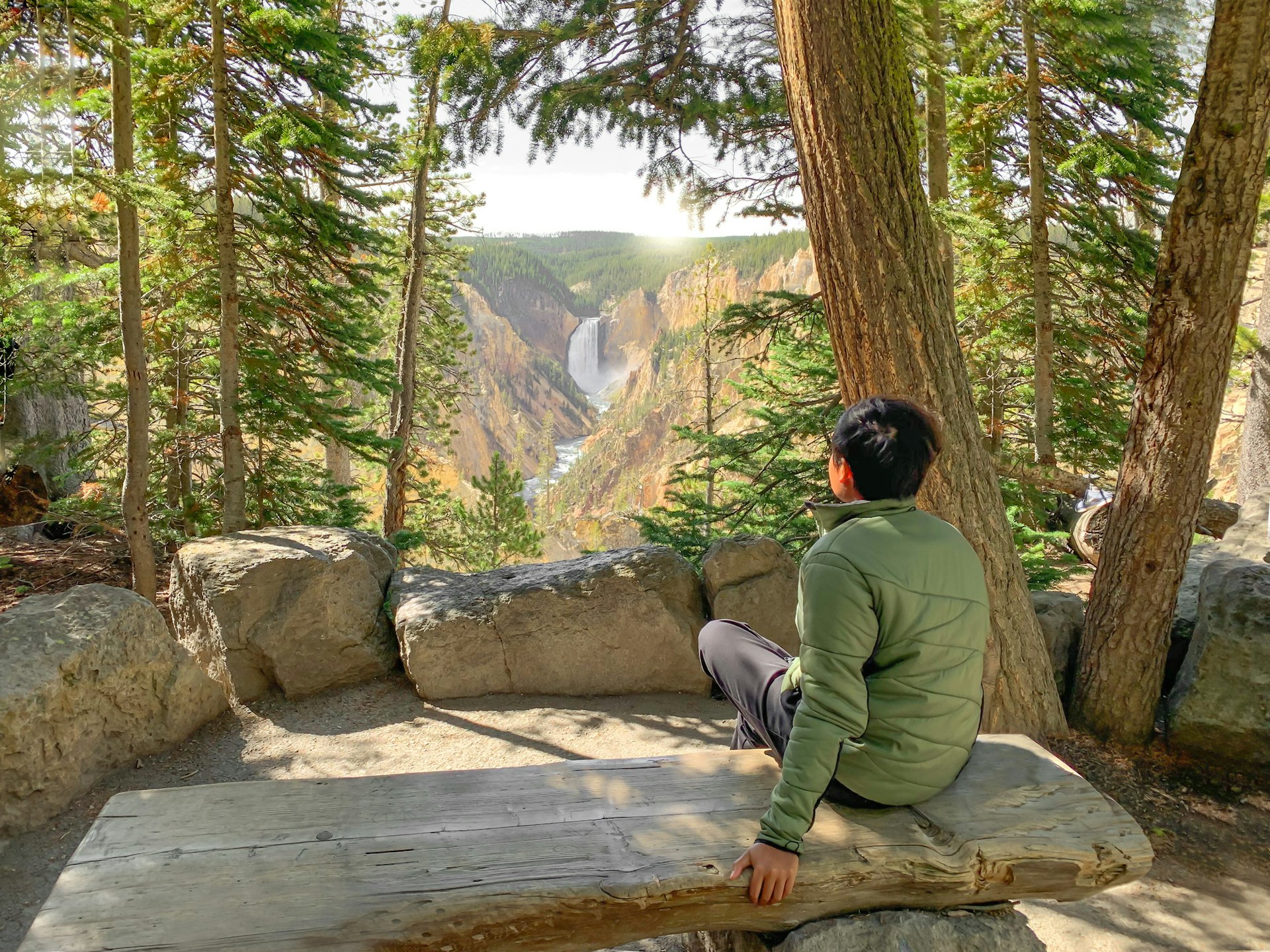 A man sits on a bench at a viewpoint and gazes towards a waterfall in the distance