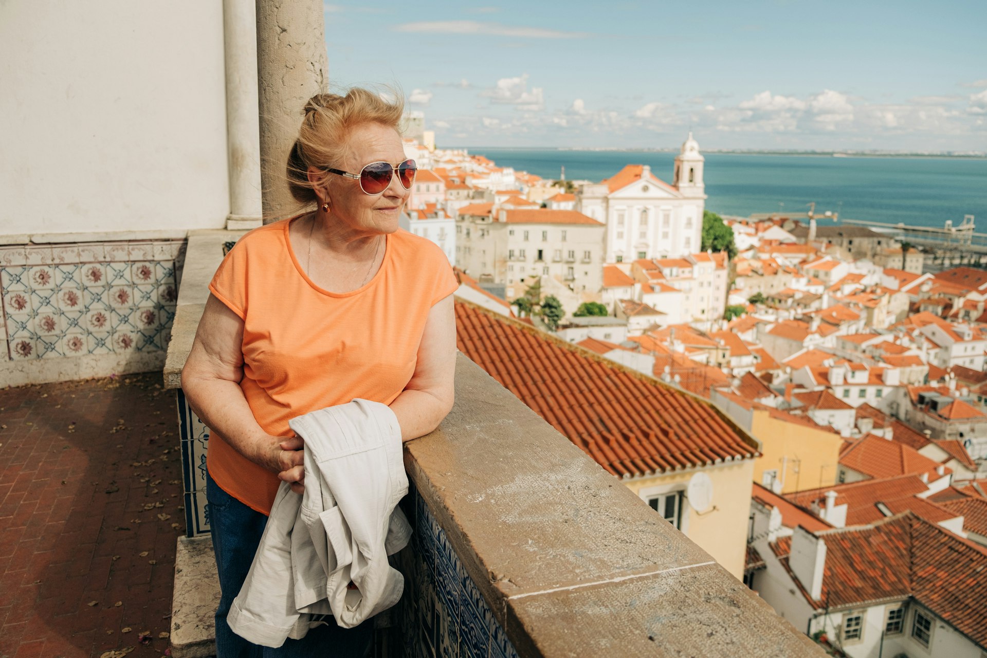 An older lady stands at a viewpoint looking out over the red roofs of buildings in a riverside city