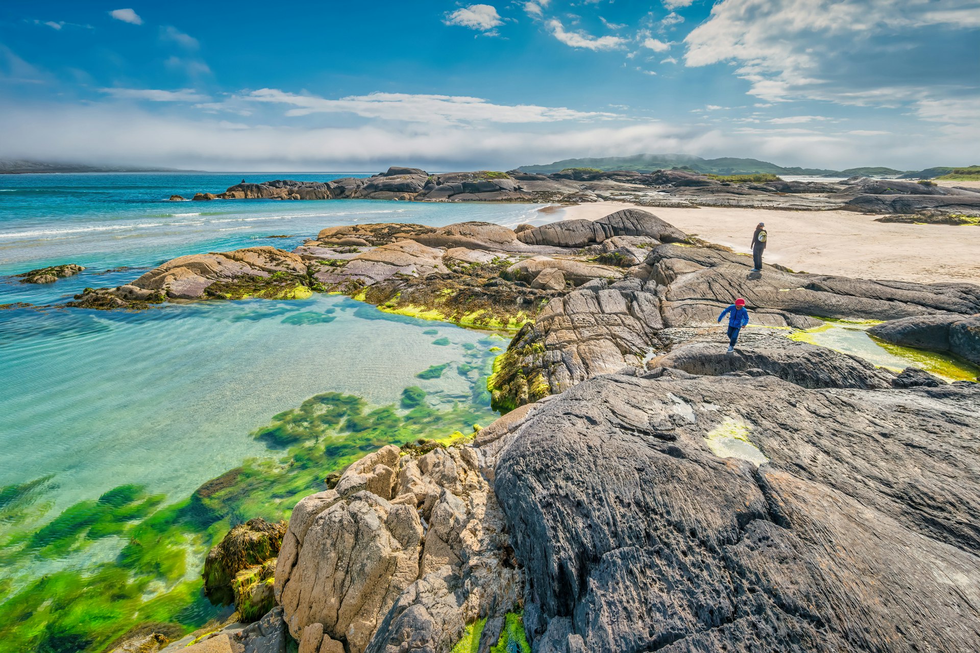 An adult and a child clamber on the rocks above a sandy beach backed by turquoise ocean in Ireland