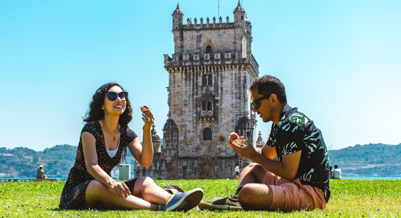 Friends sitting on grass eating a pastel de nata in front of Belem Tower in Lisbon, Portugal
1414092673