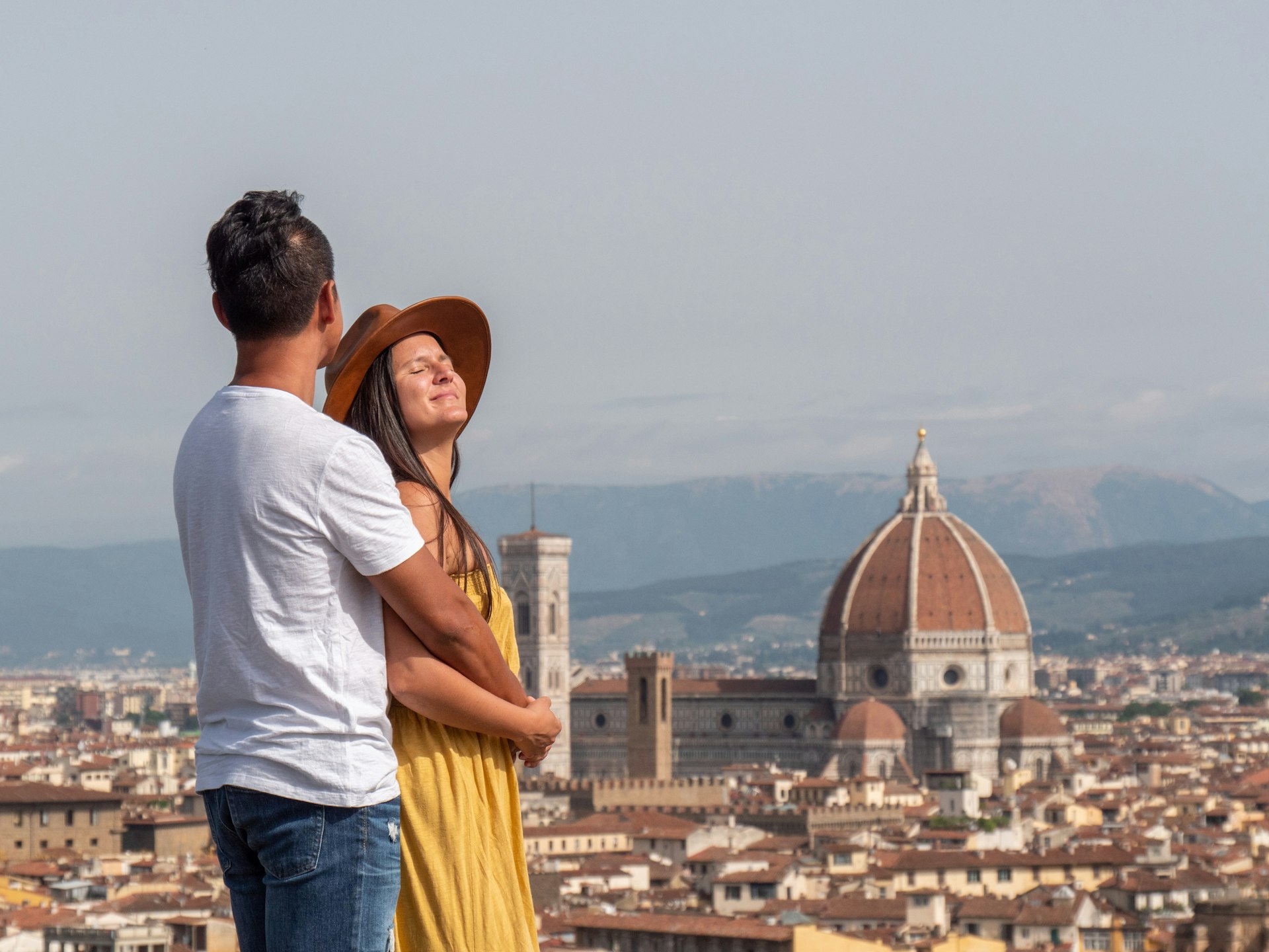 A couple, a white woman and an South Asian man, look out across the rooftops of Florence whilst in a loving embrace