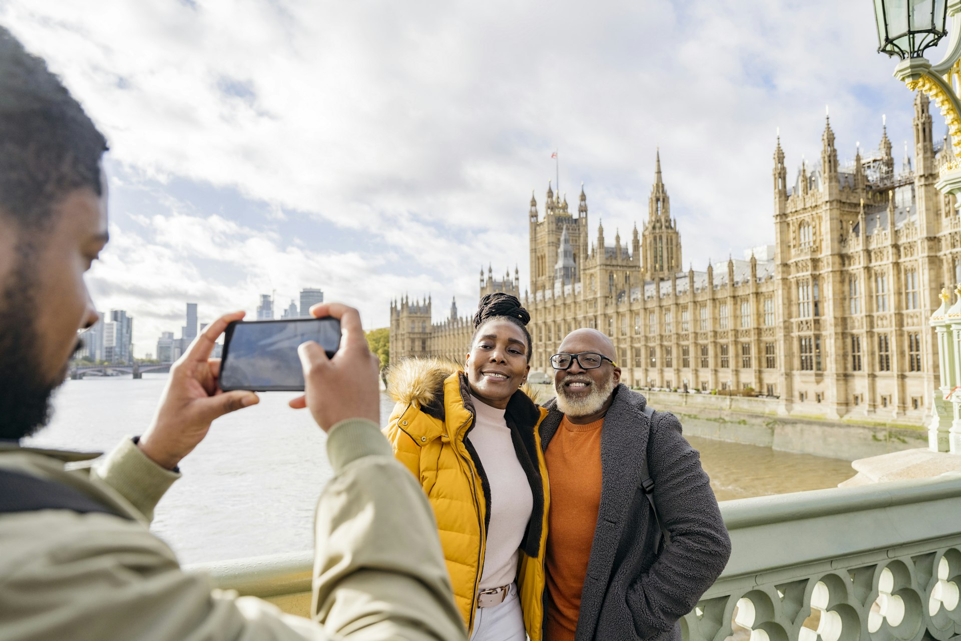 Young Black man holding smart phone and photographing mature couple in warm clothing with River Thames and Houses of Parliament in background.