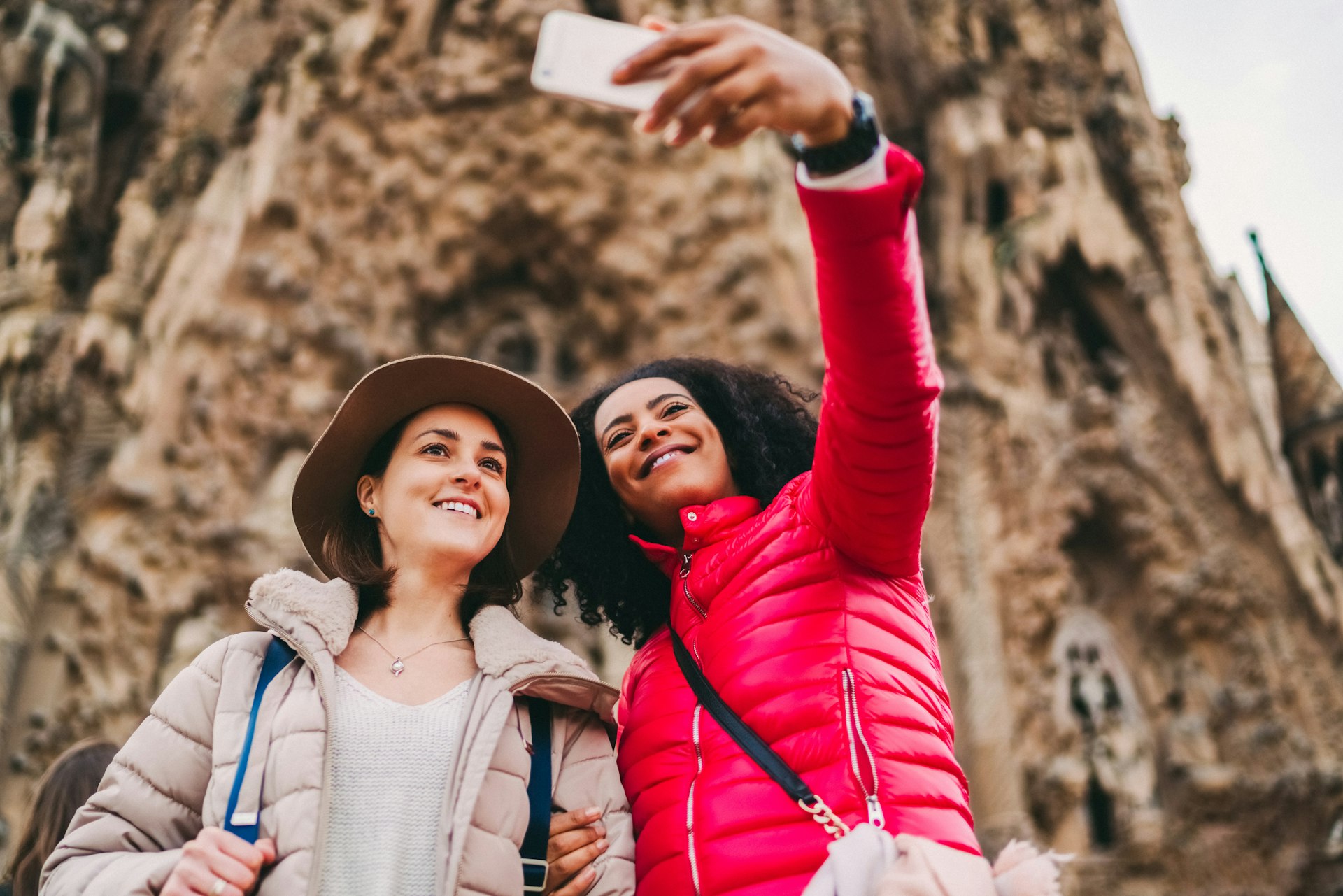 Two women stand in front of La Sagrada Familia in Barcelona taking selfies with smartphone