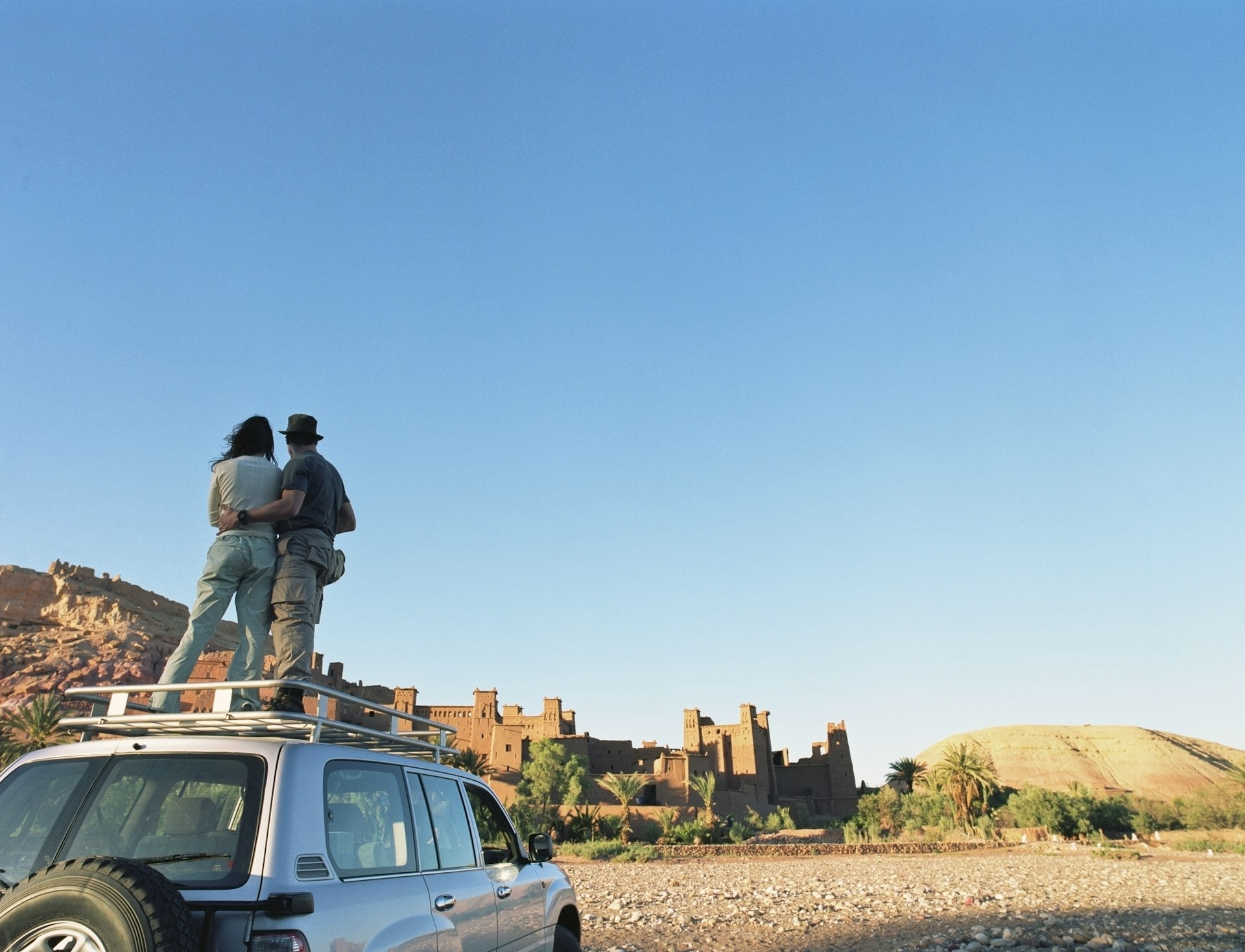 A man and woman stand hugging on the top of a four-wheel drive vehicle looking towards a red-stone fort on a hill