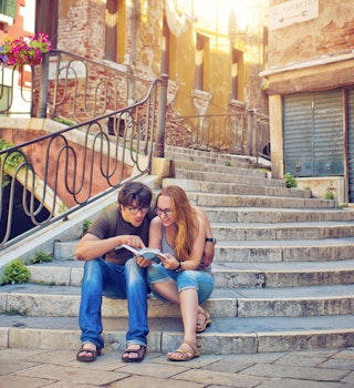 Young couple reading a guide book in Venice, Italy
506517378
Couple - Relationship, Serene People, Travel, People Traveling, Tourism, Building Exterior, Women, Men, Two People, City Life, Downtown District, Facade, Smiling, Sitting, Reading, Looking, Showing, Indigenous Culture, Heat - Temperature, History, Guidance, Journey, Happiness, Love, Adventure, Exploration, Tranquil Scene, Ancient, Old, Cultures, Famous Place, Architecture, Travel Destinations, Vacations, Urban Scene, Outdoors, Cheerful, Tourist, People, Venice - Italy, Italy, Europe, Flower, Sunlight, Summer, Sun, Staircase, House, Store, Canal, Street, Bridge - Man Made Structure, Built Structure, Cityscape, City, Town, Book
A young couple sits on steps next to a bridge in Venice and read a guidebook