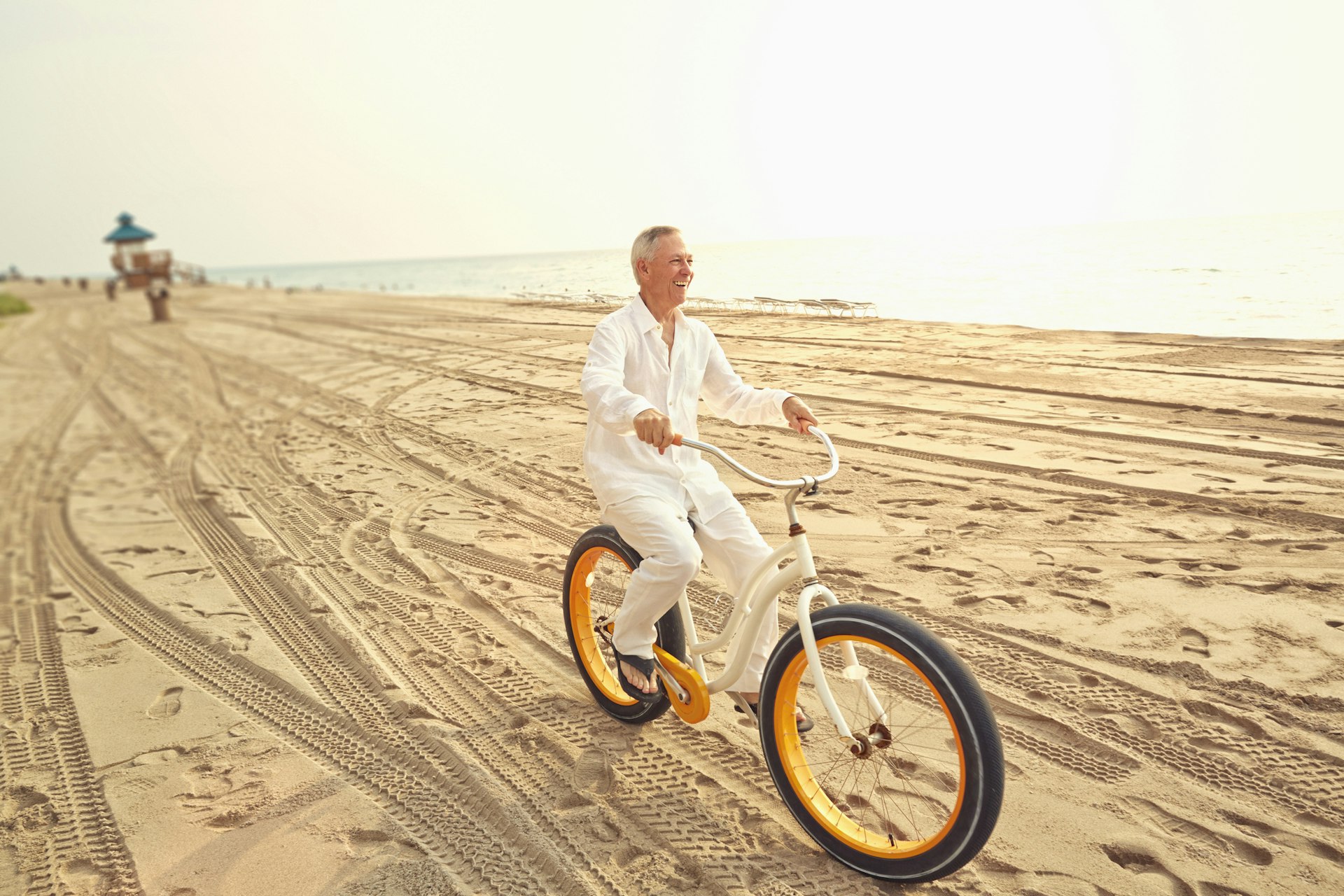 A smiling white old man with grey hair wearing all white cycles along a golden beach in Miami, US
