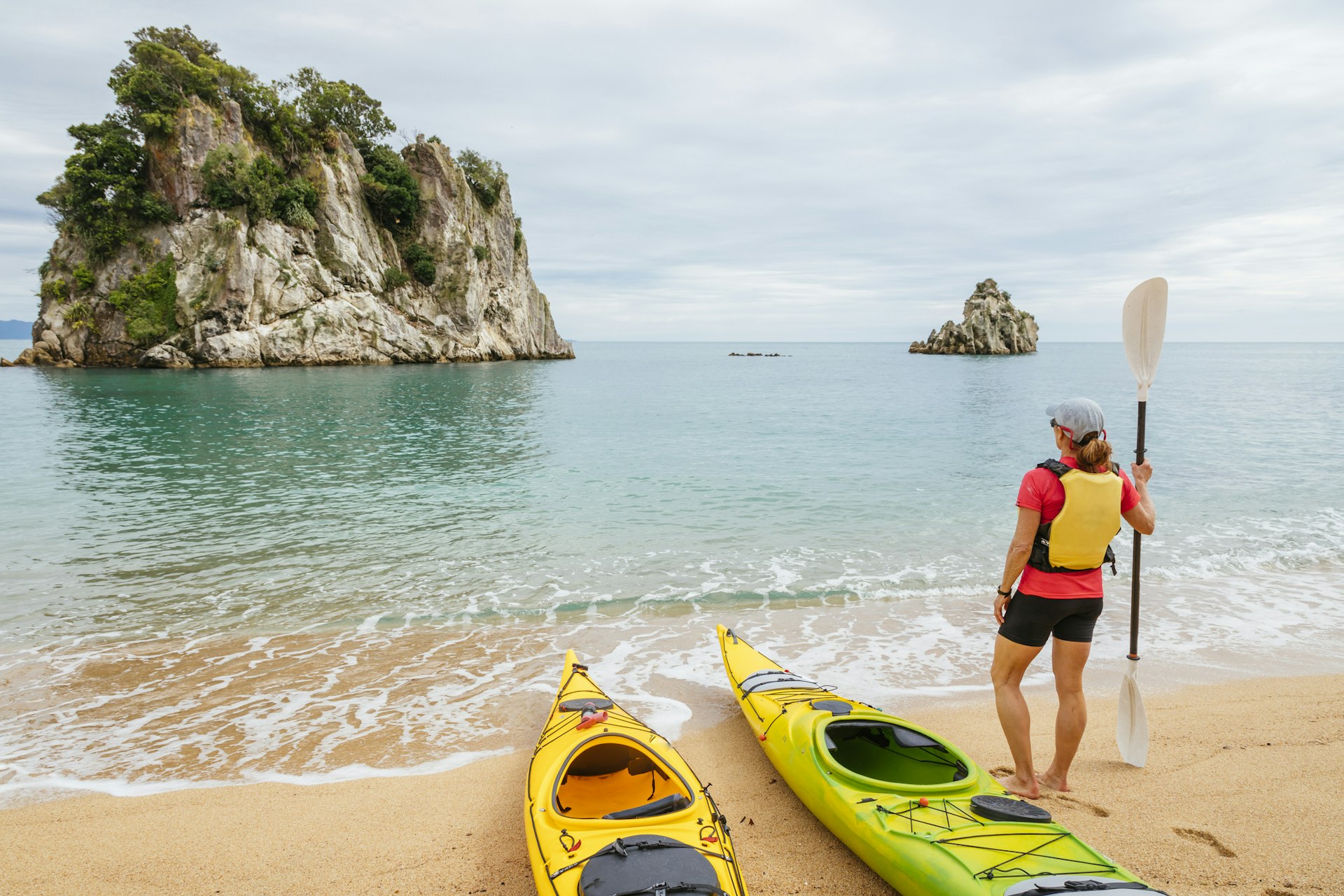 A woman stands by a yellow kayak looking out to a rocky outcrop in the sea