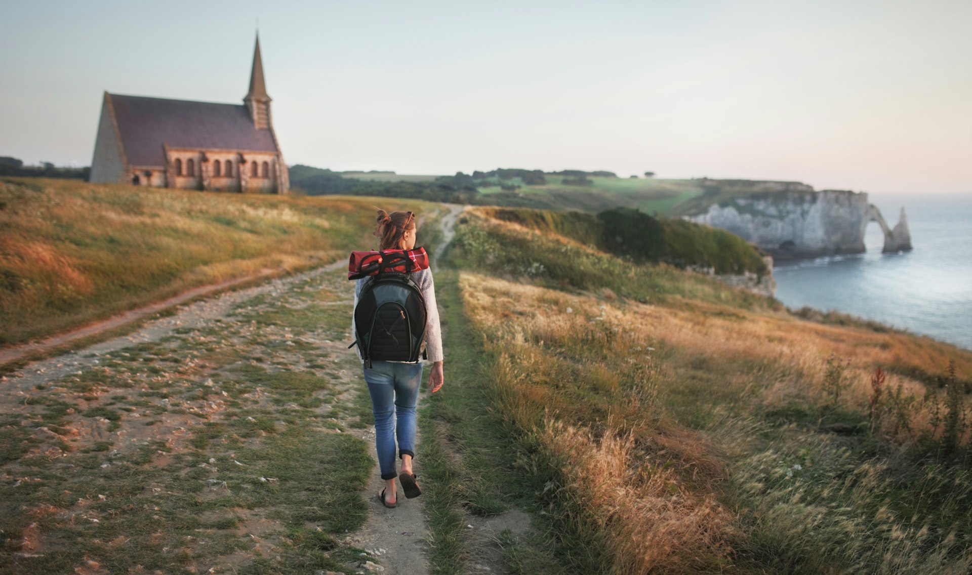 A hiker with a backpack walks along a clifftop path looking out to sea on her right. To her left is a small church with a spire.