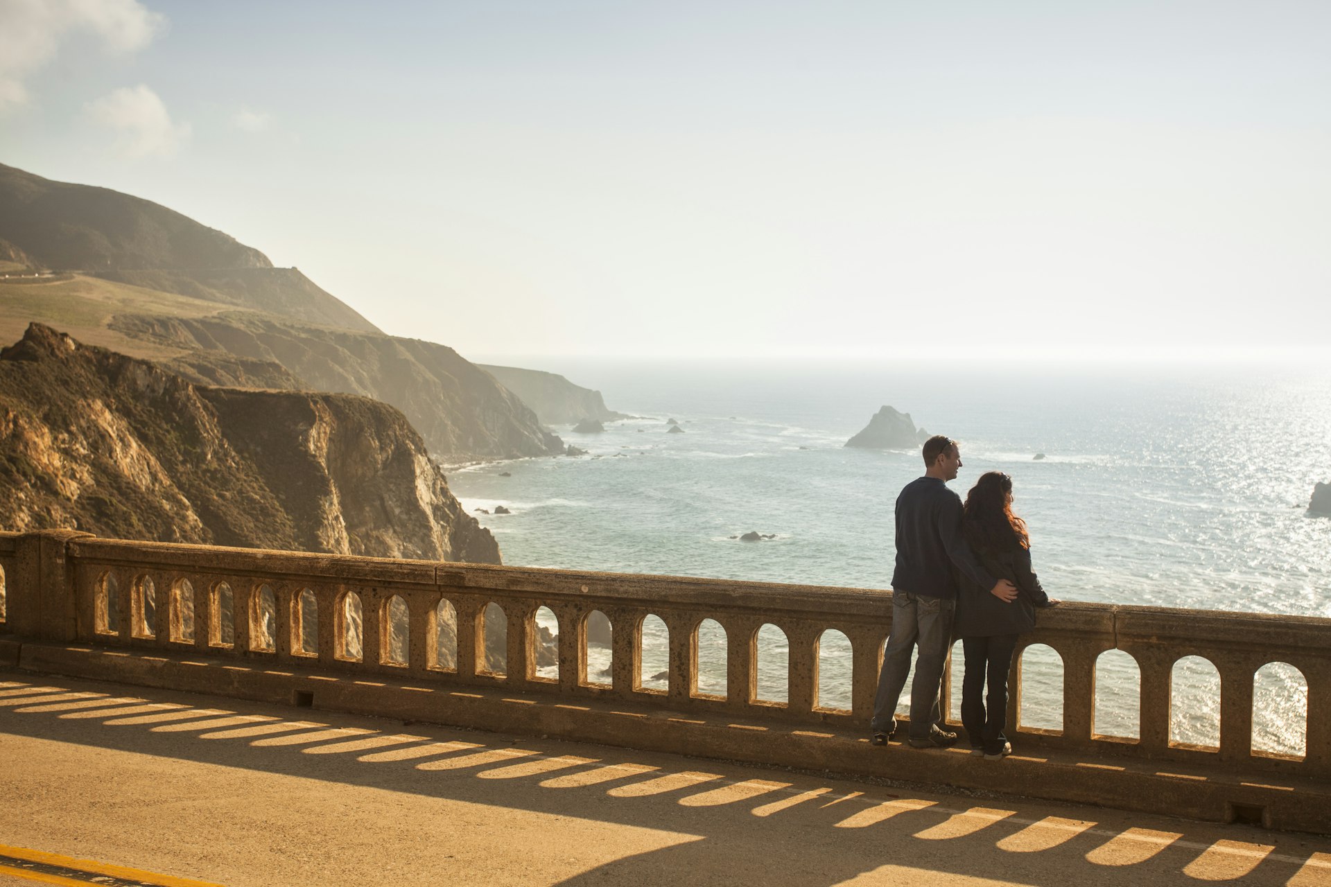 A couple standing on the Bixby Bridge, looking over the view, on highway 1 near the rocky Big Sur coastline of the Pacific Ocean California, USA