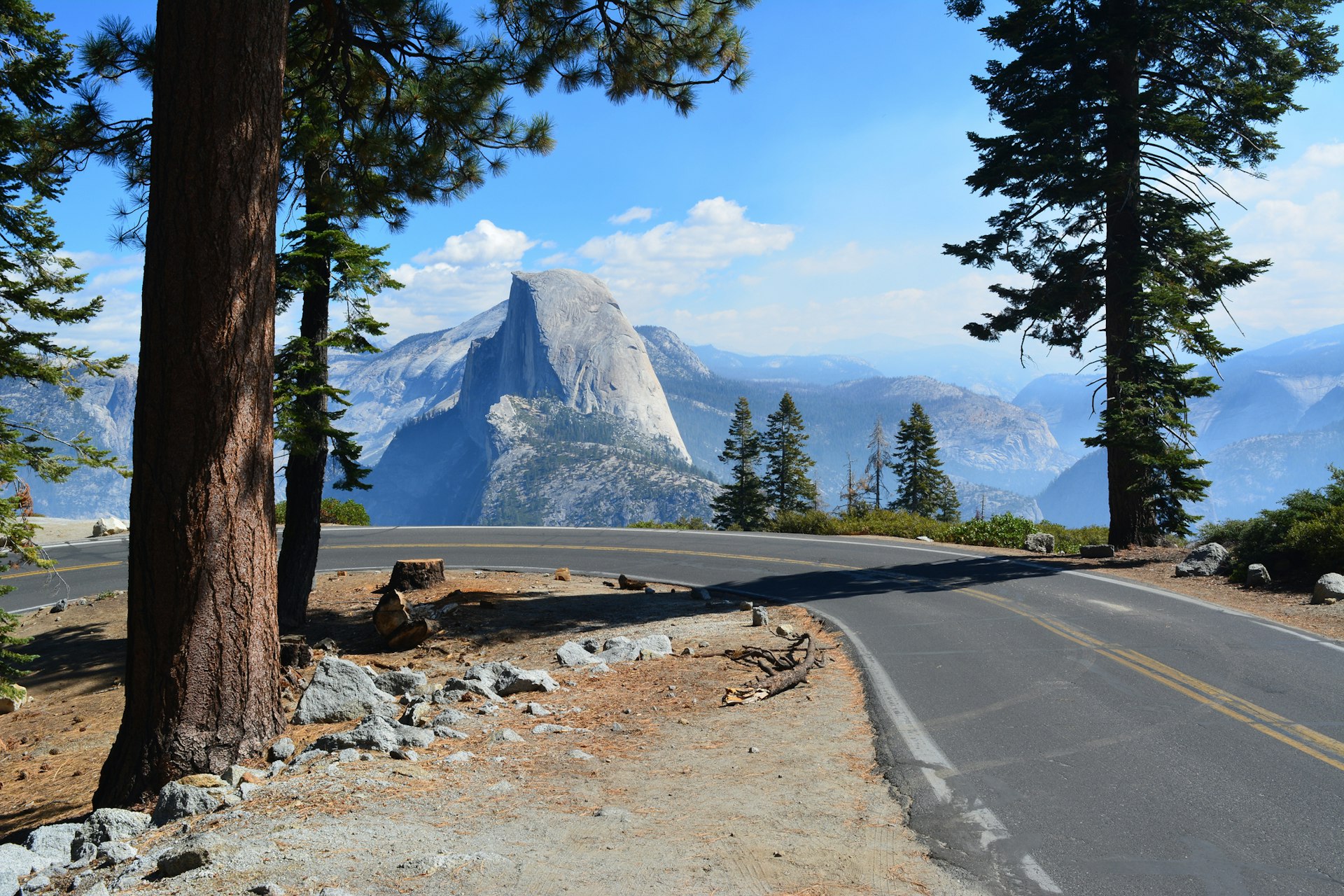 A road bends on the edge of a viewpoint looking towards Yosemite's famous Half Dome, a huge rock structure in the shape of a dome cut in half