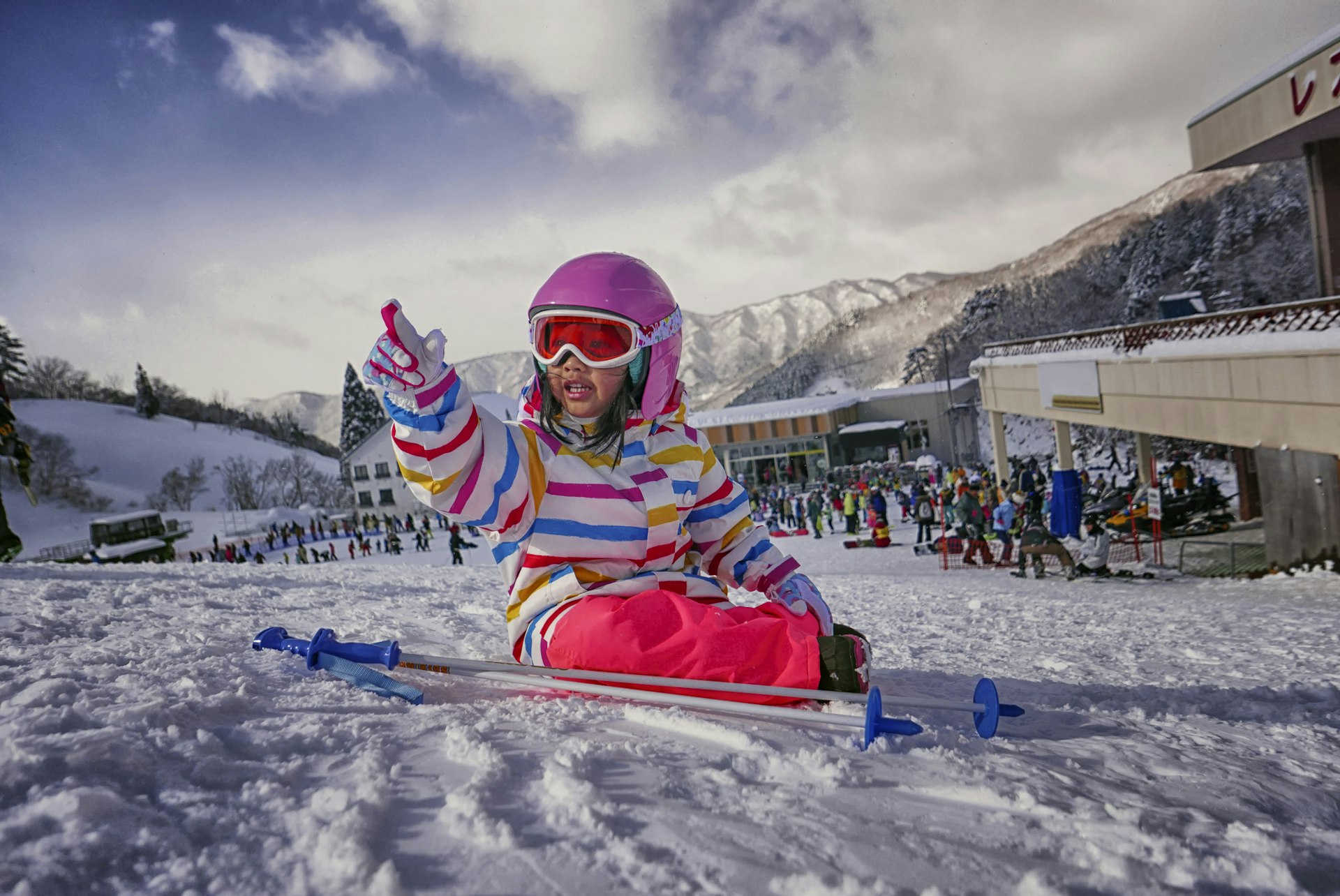 A small Japanese child sits playing in the snow on a ski slope