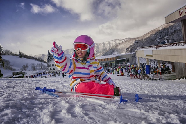 A small Japanese child sits playing in the snow on a ski slope