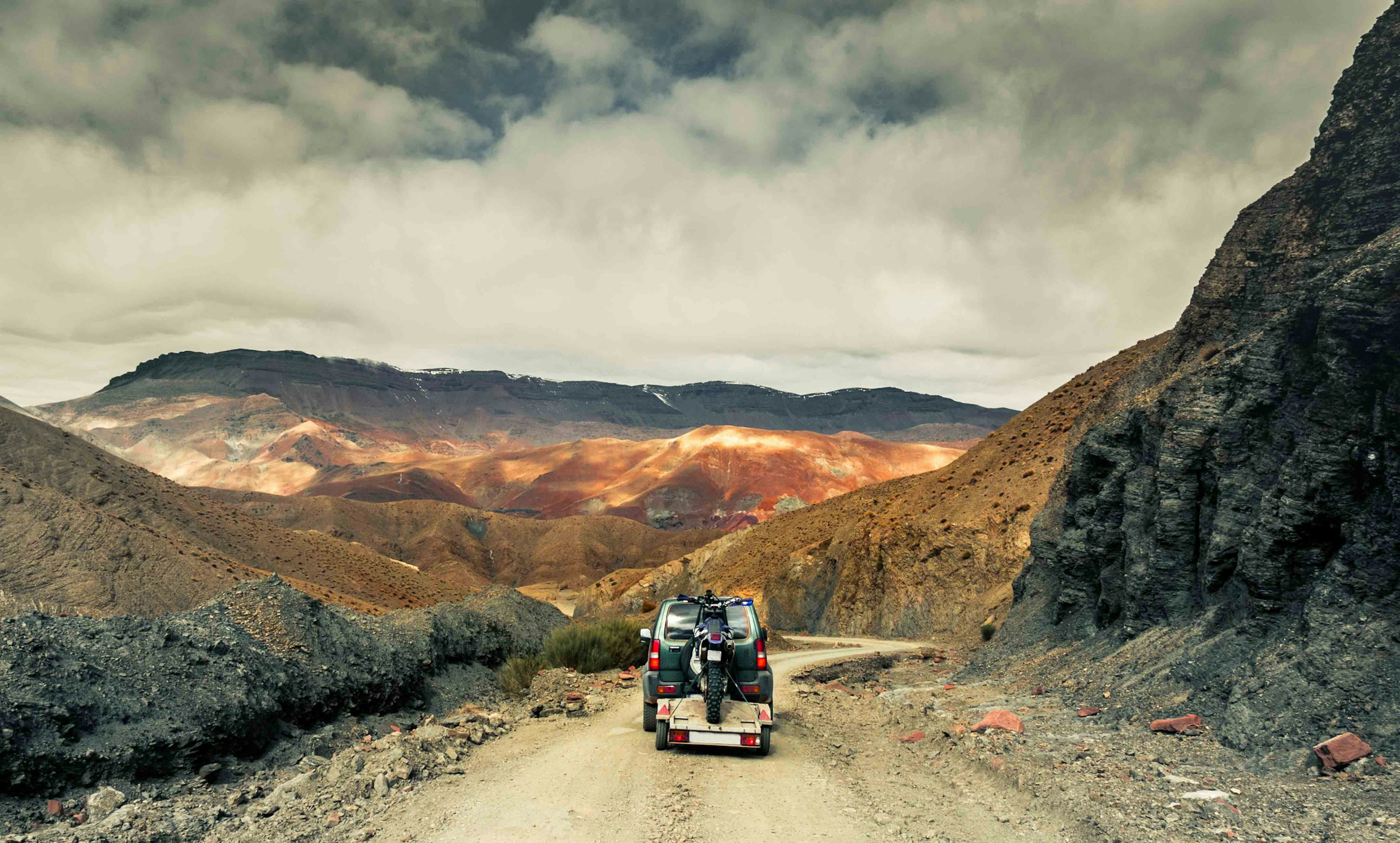 The back of a four-wheel-drive vehicle towing a trailer with a dirt bike on it on a trail in a mountain range