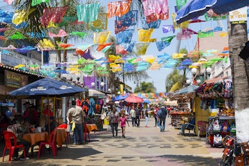 People shop and walk below colorful hanging flags at Plaza Santa Cecilia, a historic Mexican square in the heart of the city.