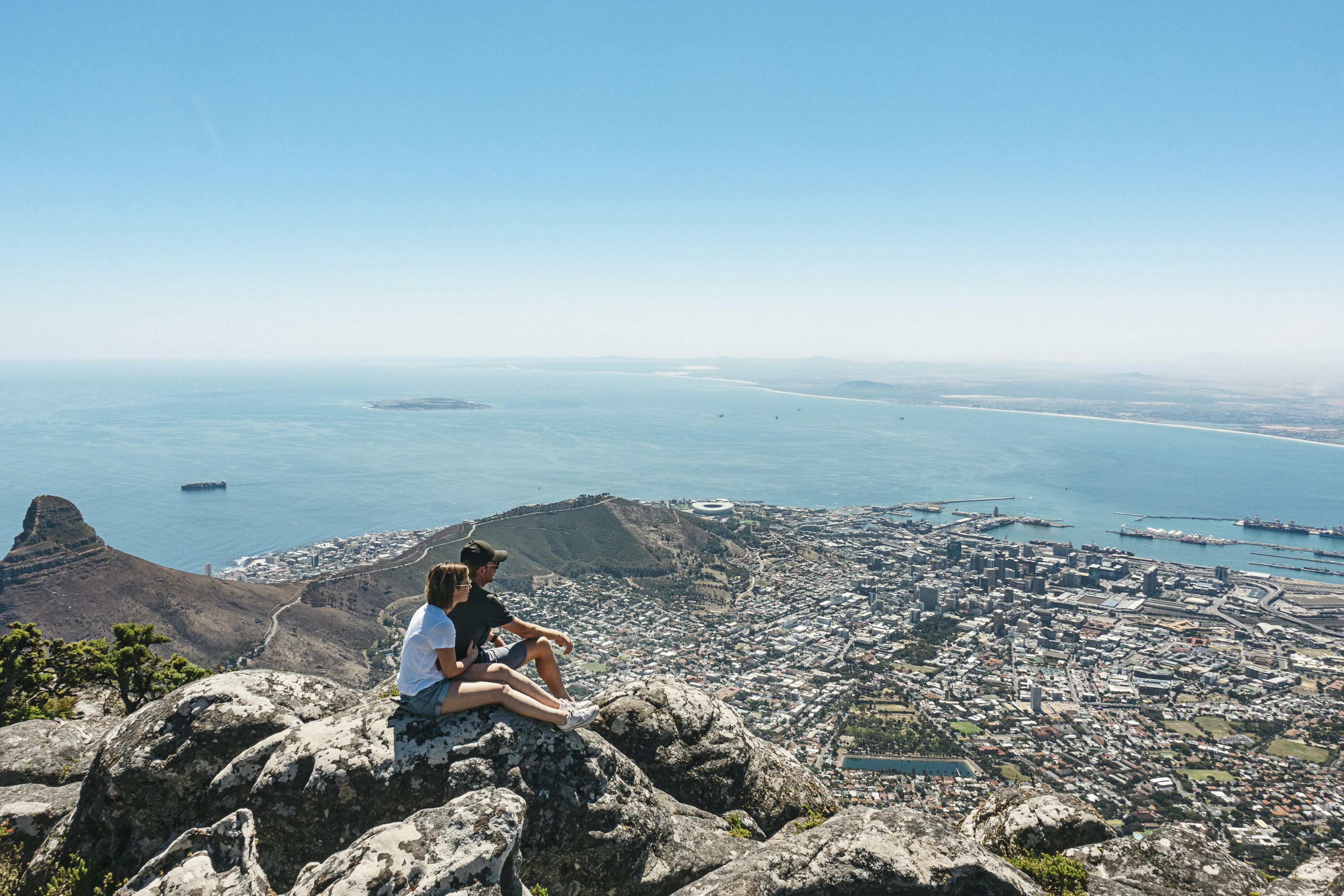 A couple sit on a rock overlooking a sprawling city below