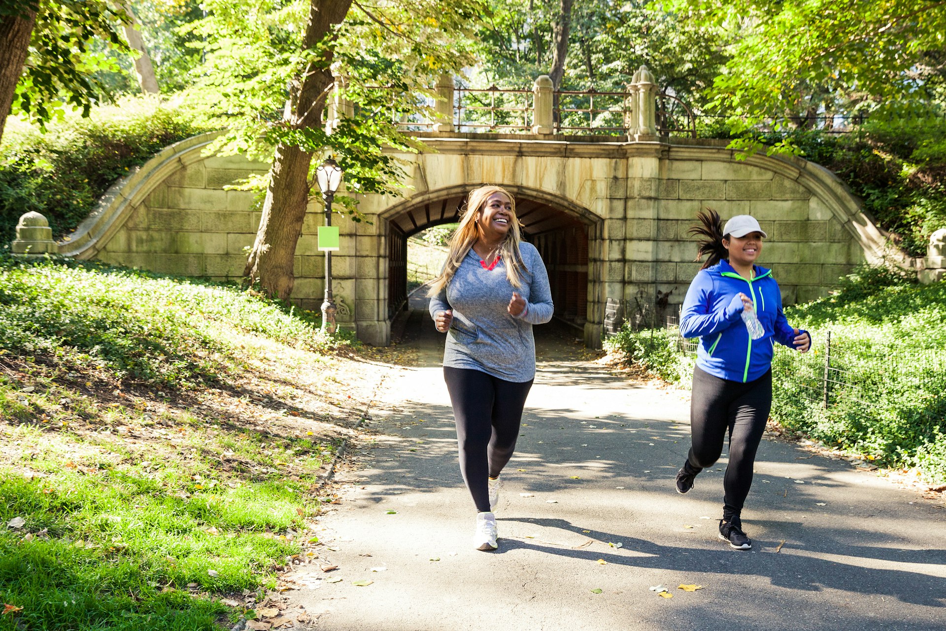 Two plus size women jogging in Central Park on a sunny day past a bridge