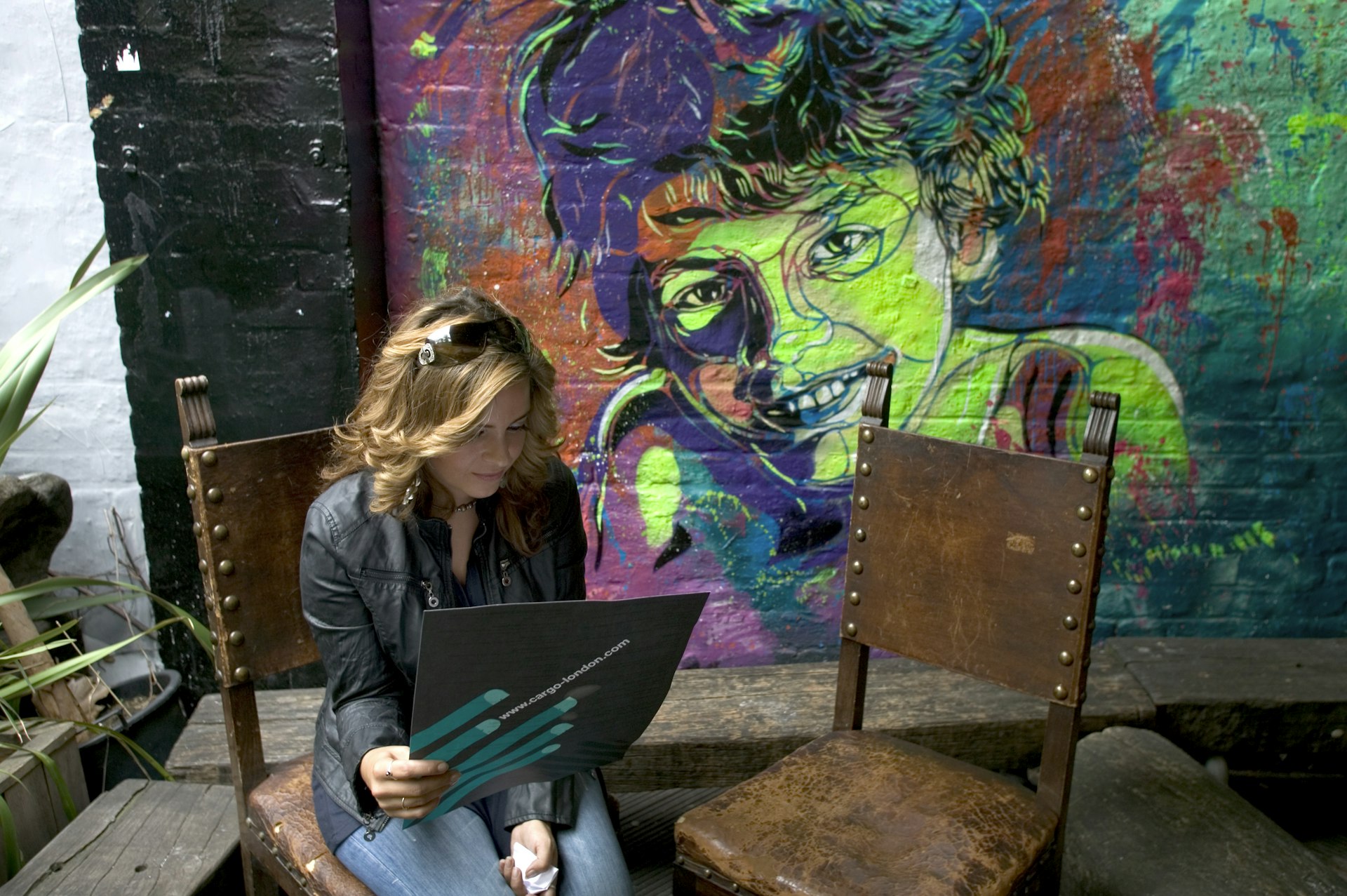 A brown hair white woman, around 40 years old, sits on an old brown chair next to an empty chair with street art behind her of a woman in green and purple taken inside Cargo Club, Shoreditch 