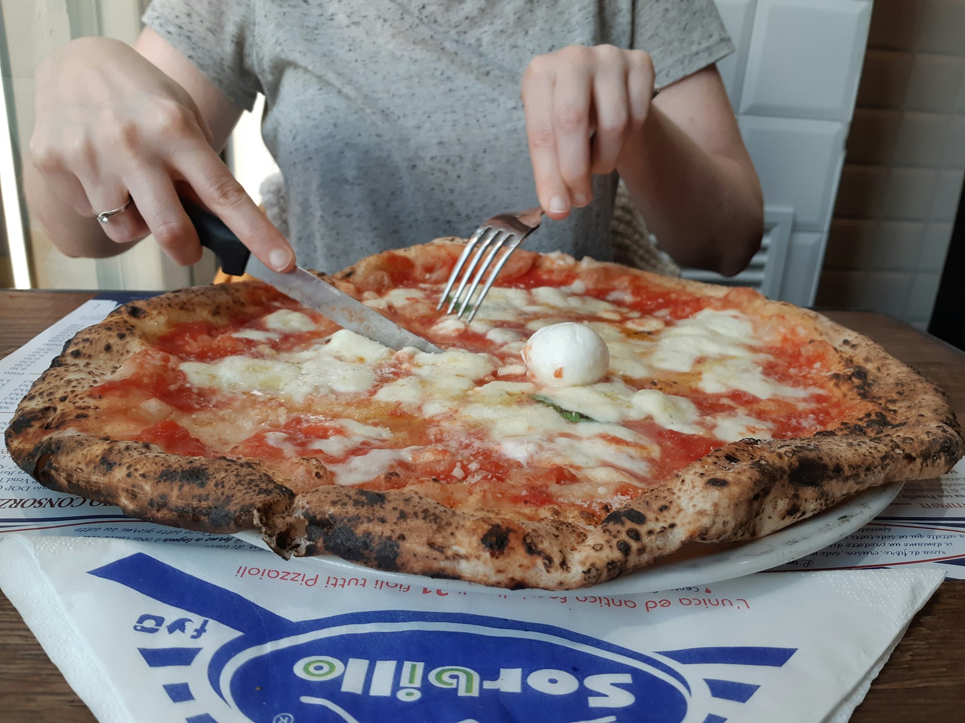 A person sits at a table with a large pizza on it in a blue-and-white box