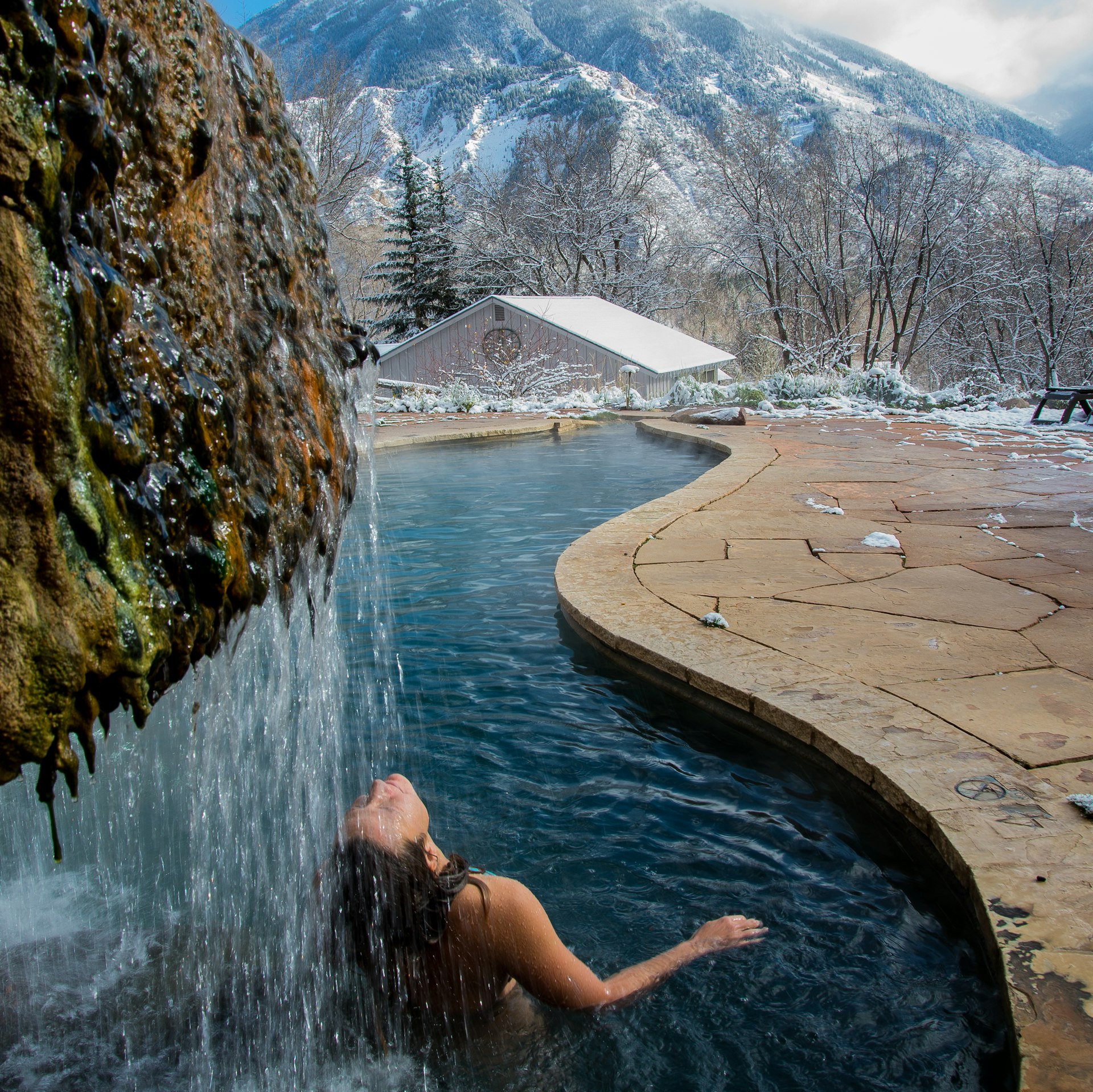 A woman leans back into a waterfall at a natural hot spring at Avalanche Ranch Hot Springs, Colorado, USA