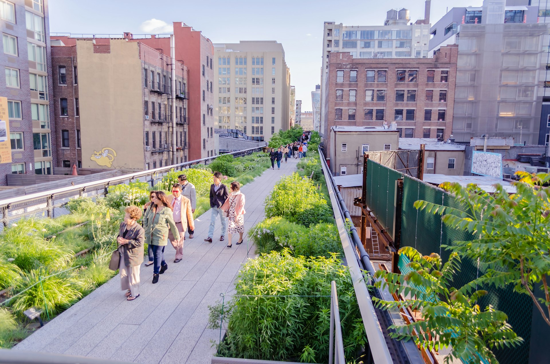 People walk along a raised boardwalk lined with flower beds and greenery surrounded by city apartments