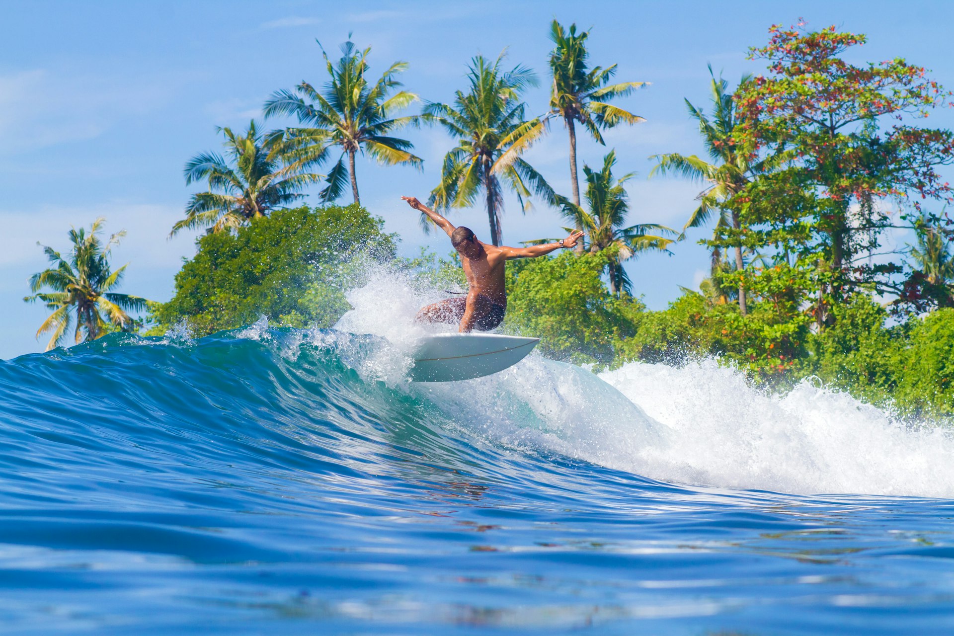 A shirtless man rides a wave in the waters around Bali, Indonesia, with tropical palm trees in the background