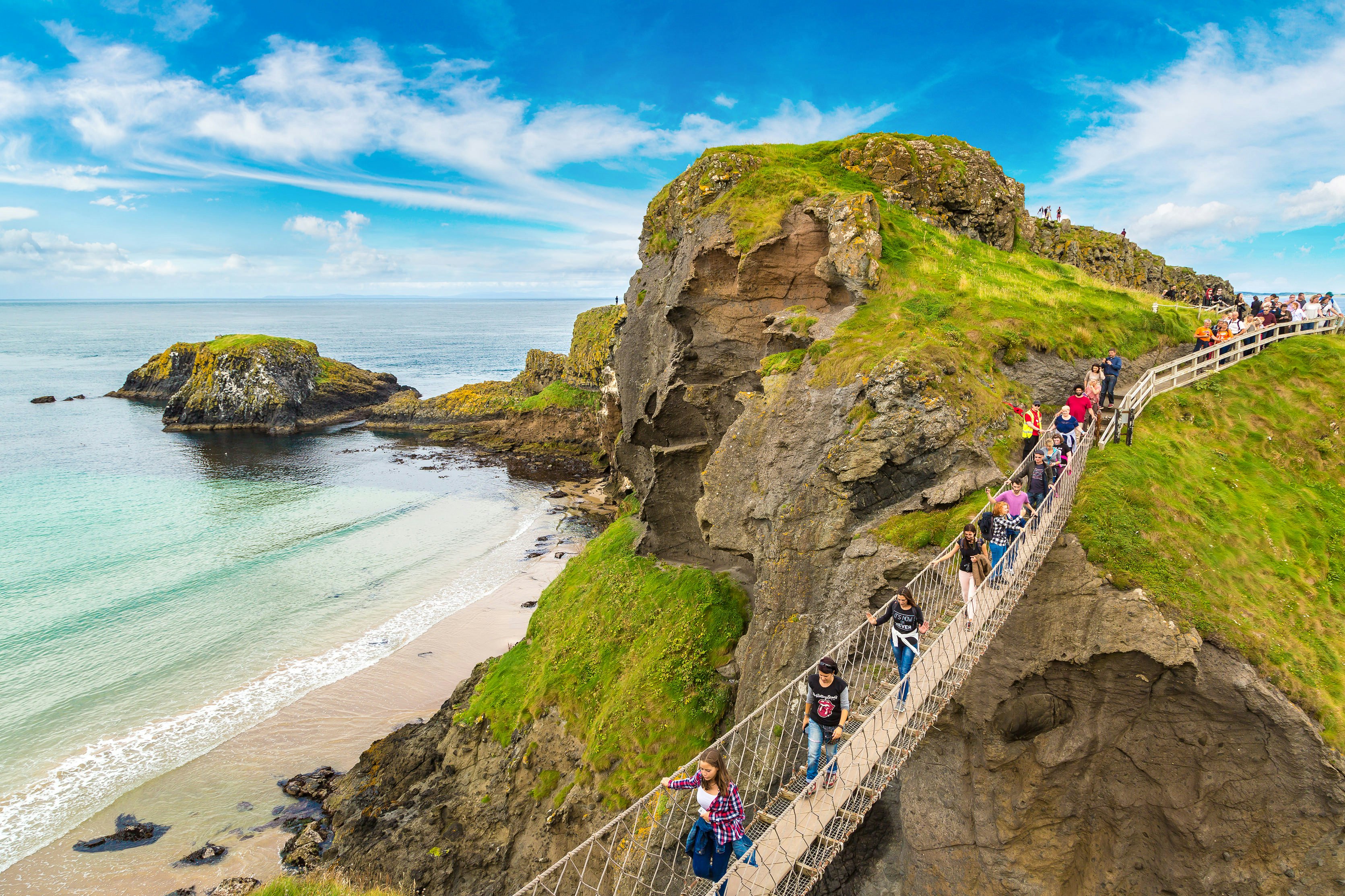An overhead view of people walking across a narrow bridge that spans a gorge above a beach by a rocky headland.