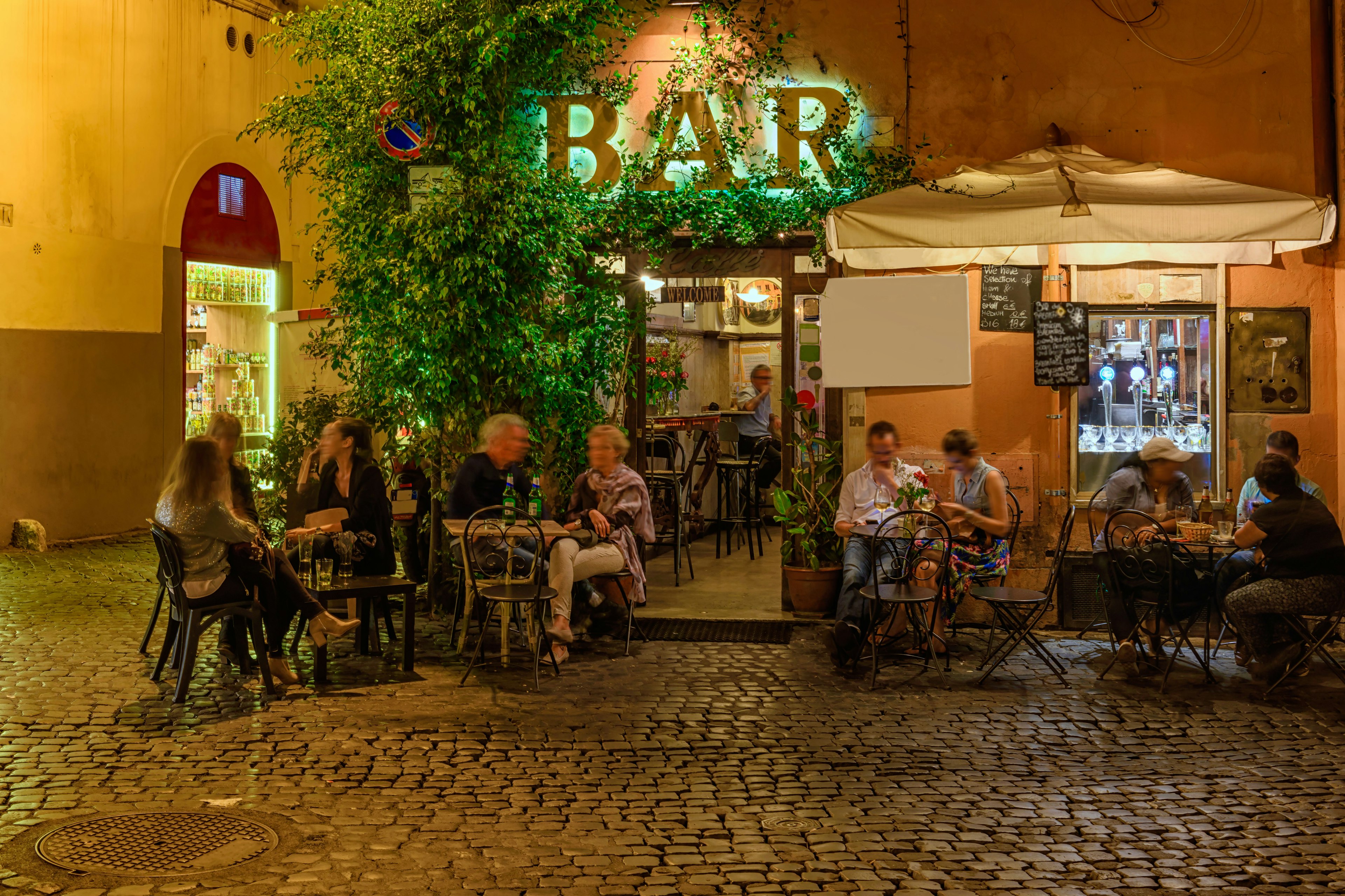 People sit outside a bar on an old street at night in Trastevere, Rome