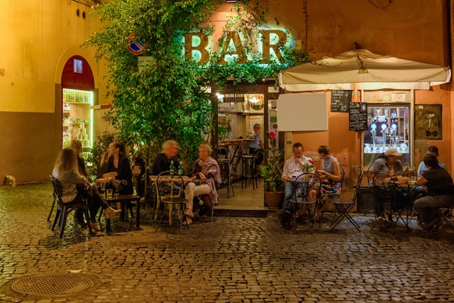 People sit outside a bar on an old street at night in Trastevere, Rome
