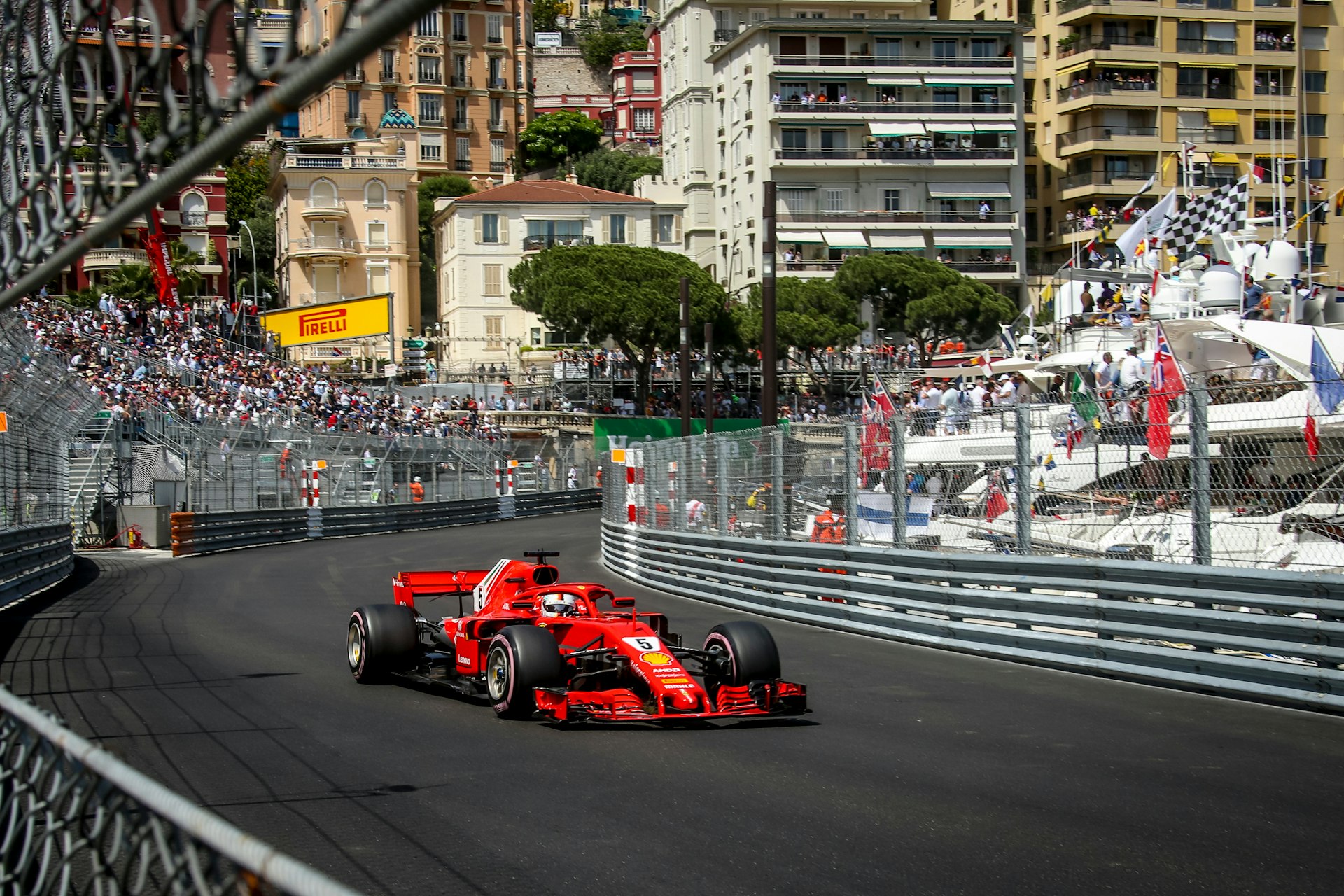 A red Formula-1 car on a track with a seating area on one side of the track and luxury yachts on the other