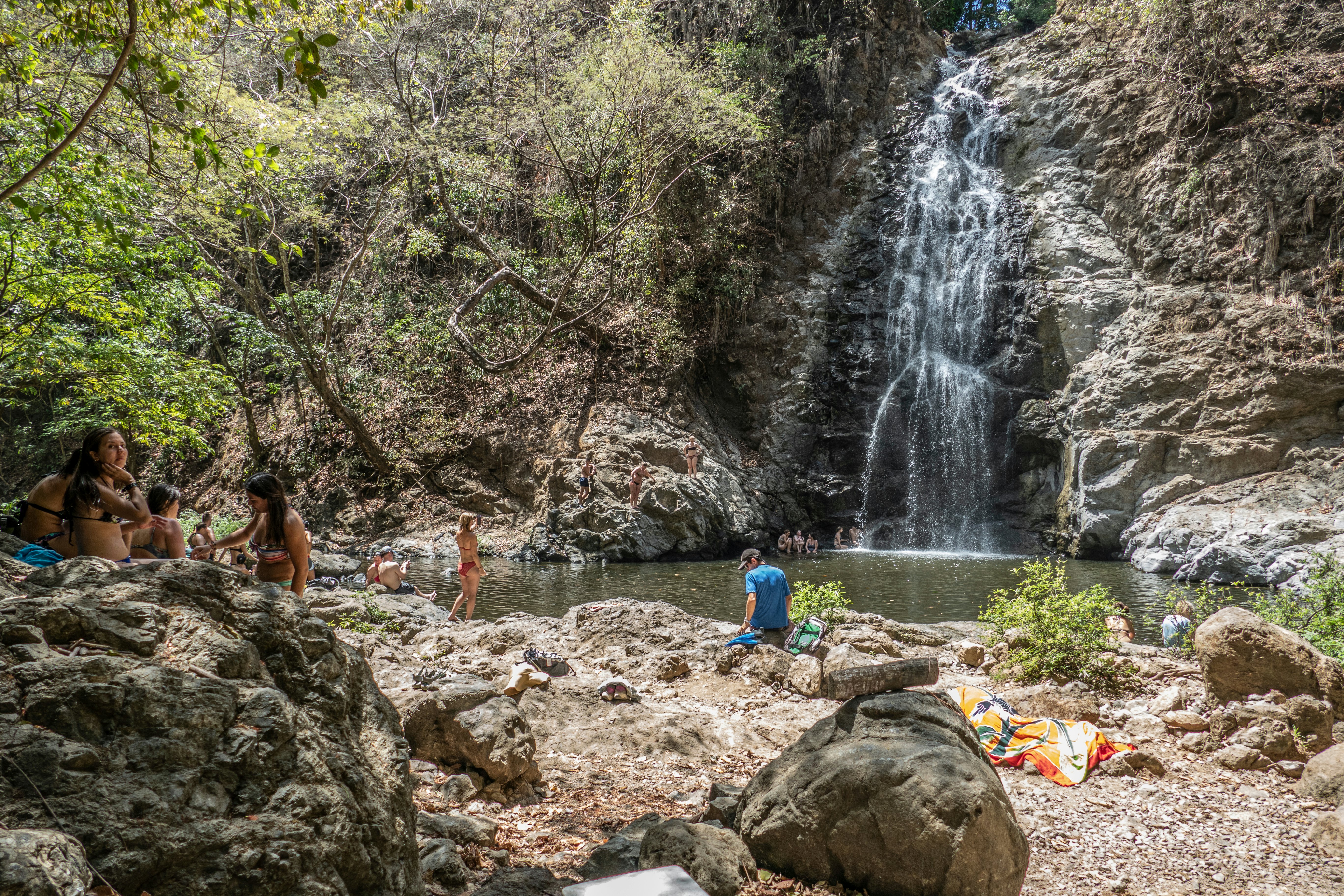 People in swimsuits are gathered on the rocks around a pool of water by a waterfall in Costa Rica.