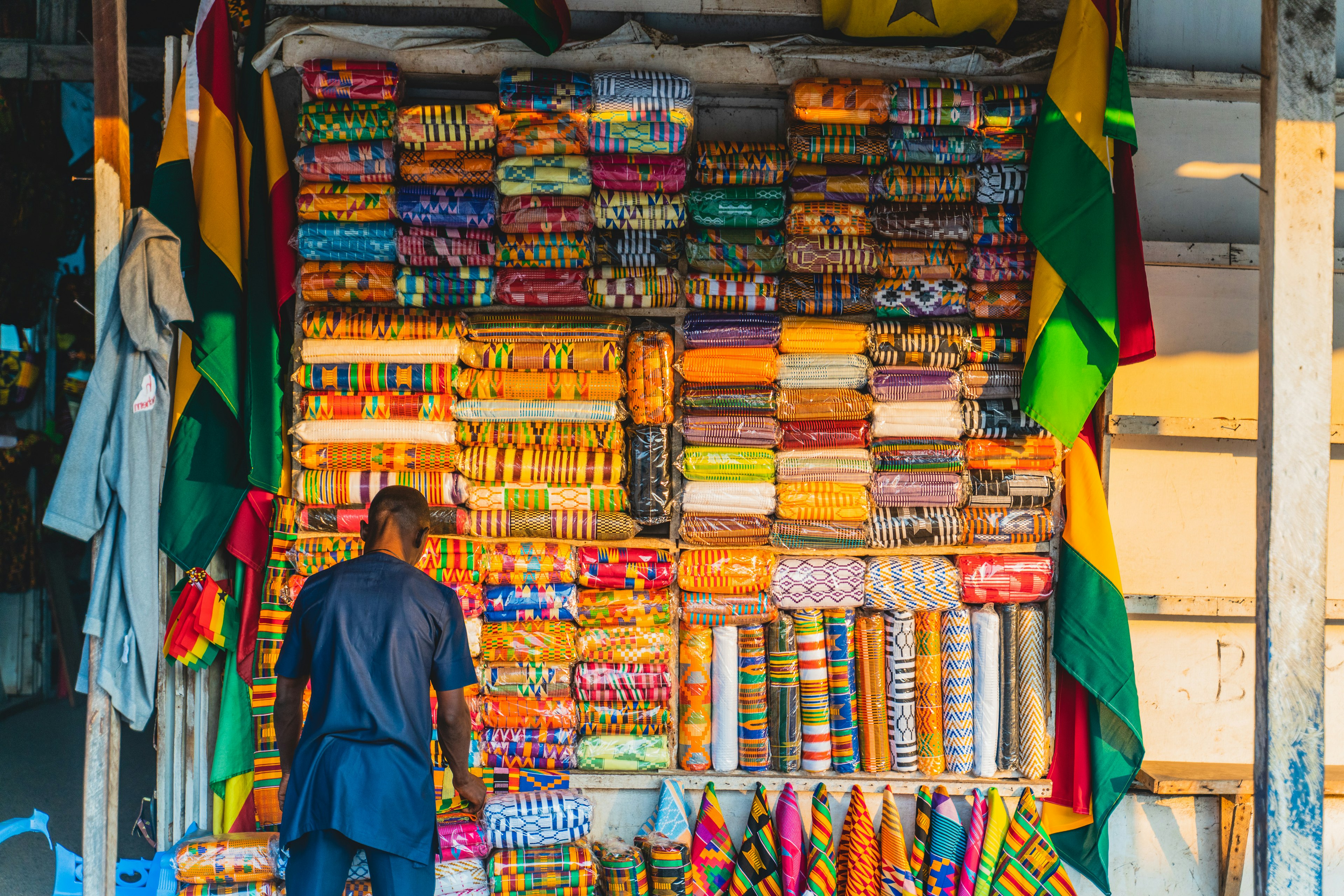 A man stands at a market stall stocked with brightly colored and heavily patterned fabrics