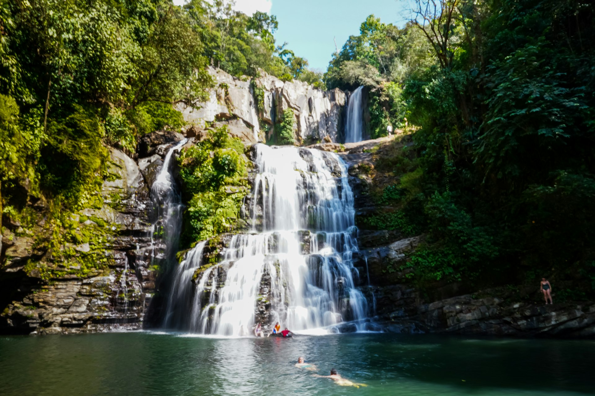 People swim and play in the water at the base of a tiered waterfall in a jungle setting