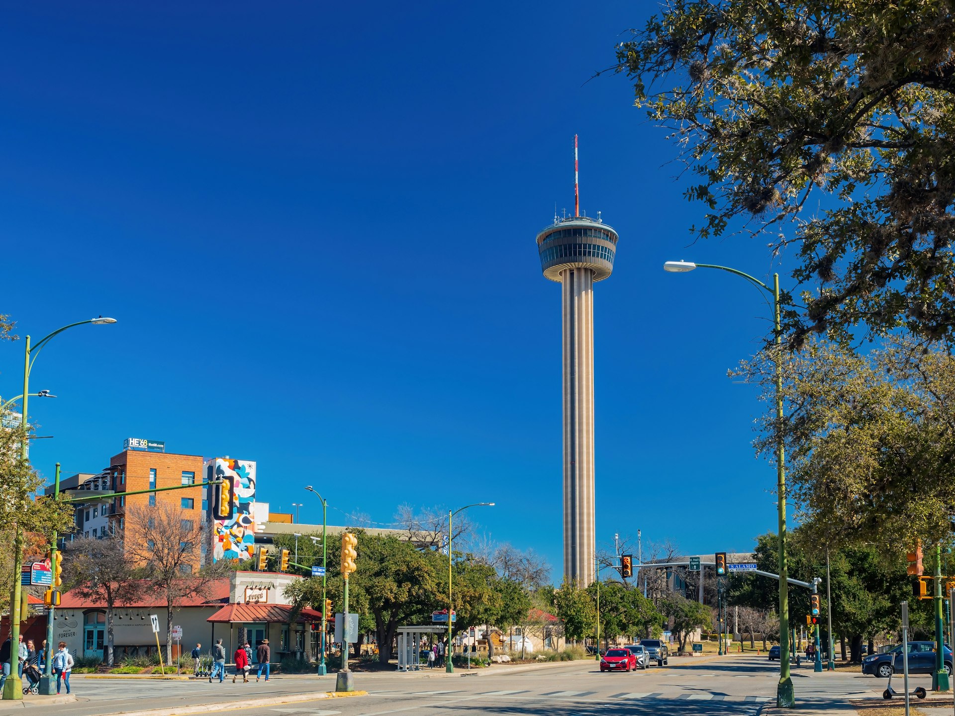 Sunny view of people walking and driving on a street in San Antonio, with the Tower of the Americas in the background 