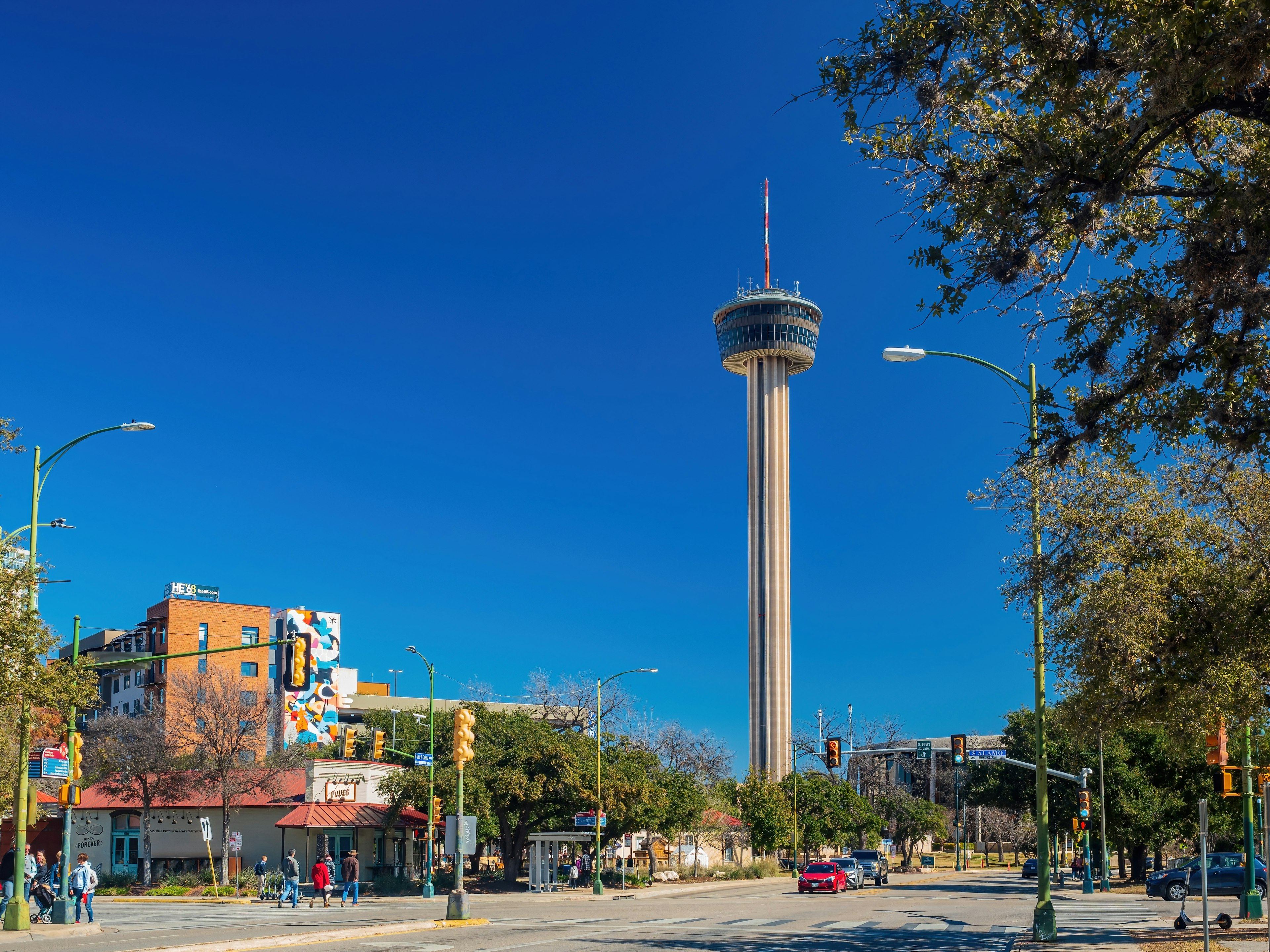 Sunny view of people walking and driving on a street in San Antonio, with the Tower of the Americas in the background