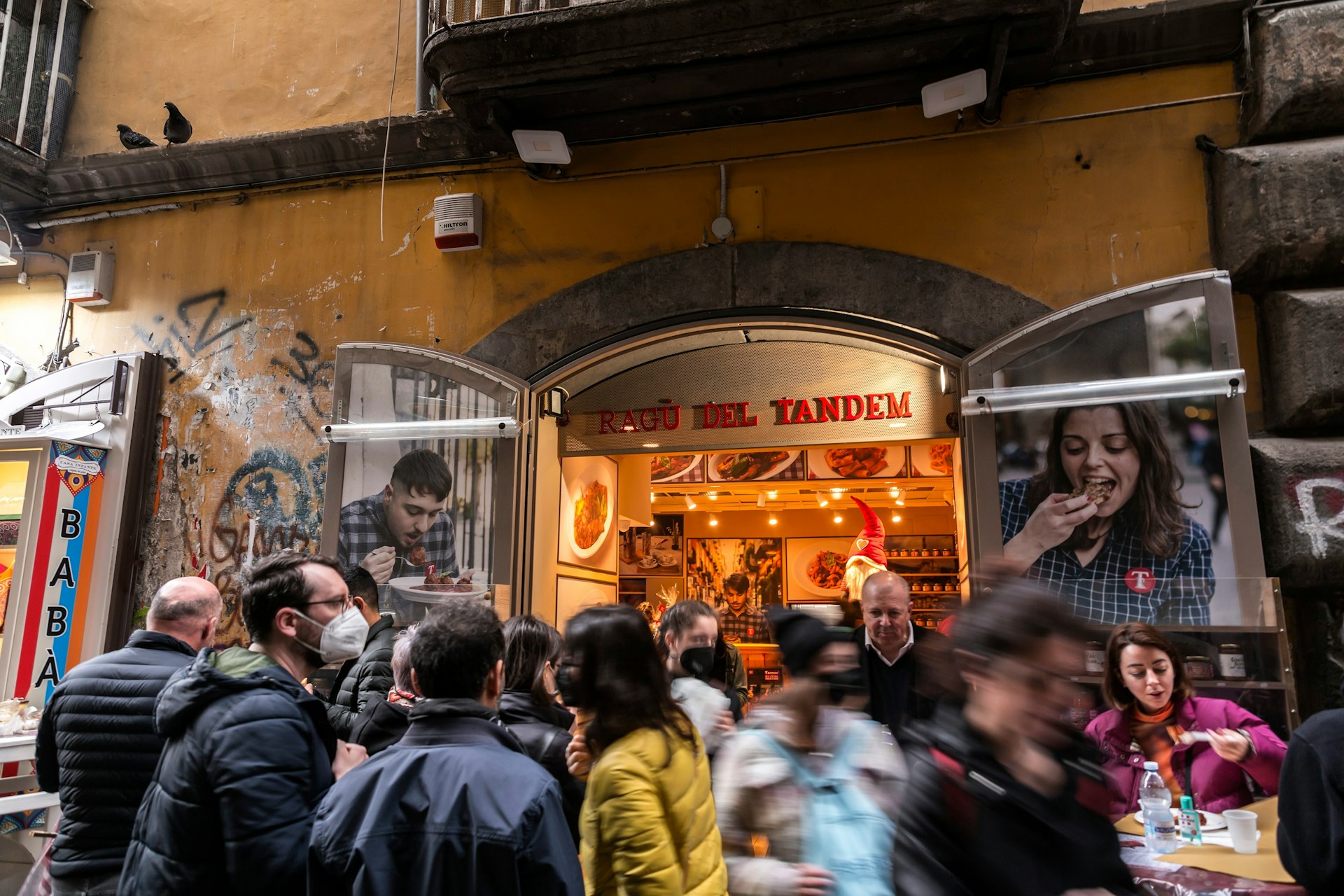 People in the street crowd around a doorway to a restaurant waiting for a table