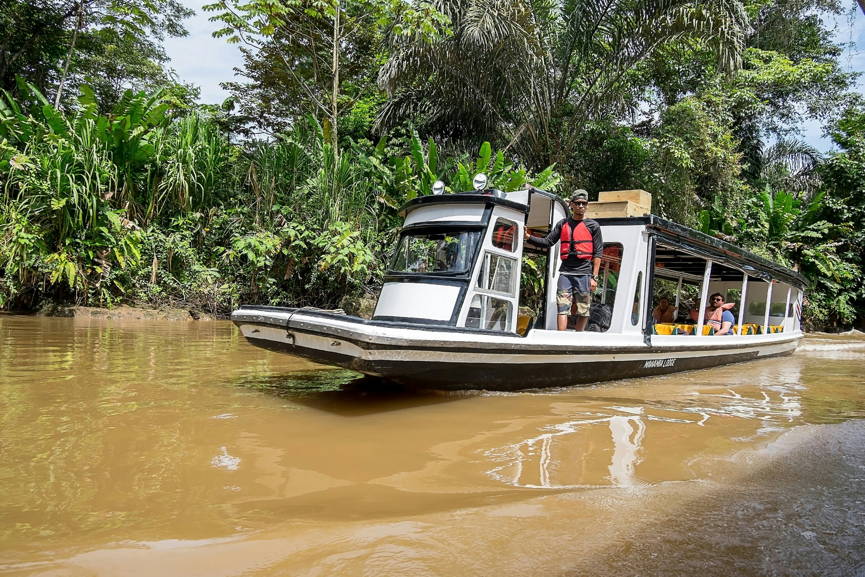 A boat with open sides navigates a muddy-looking waterway in Costa Rica; passengers are sitting inside, and a person in a red life vest stands on the deck at the front.