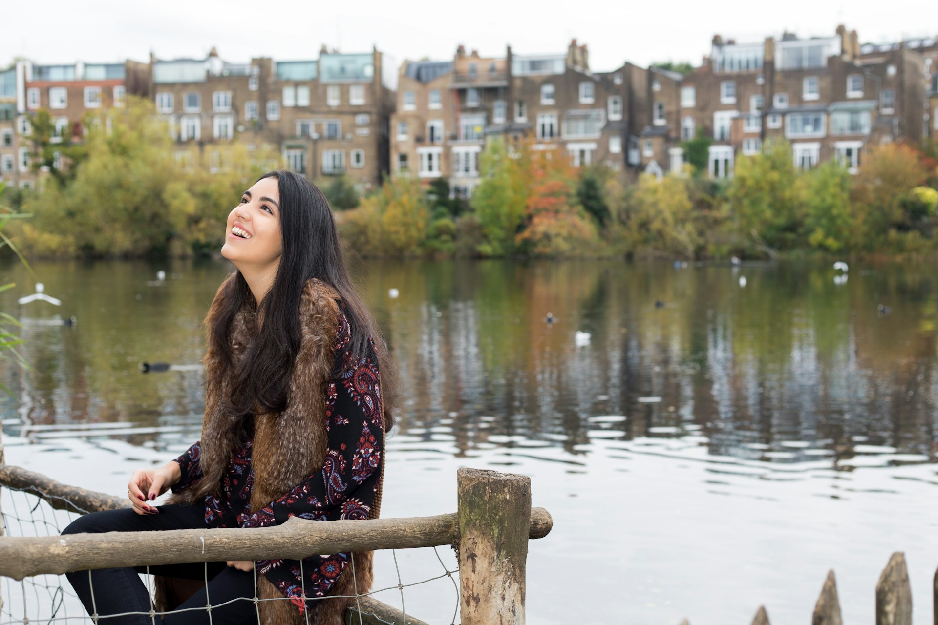 Young white woman with dark brown hair relaxes by Hampstead Heath lake in the autumn