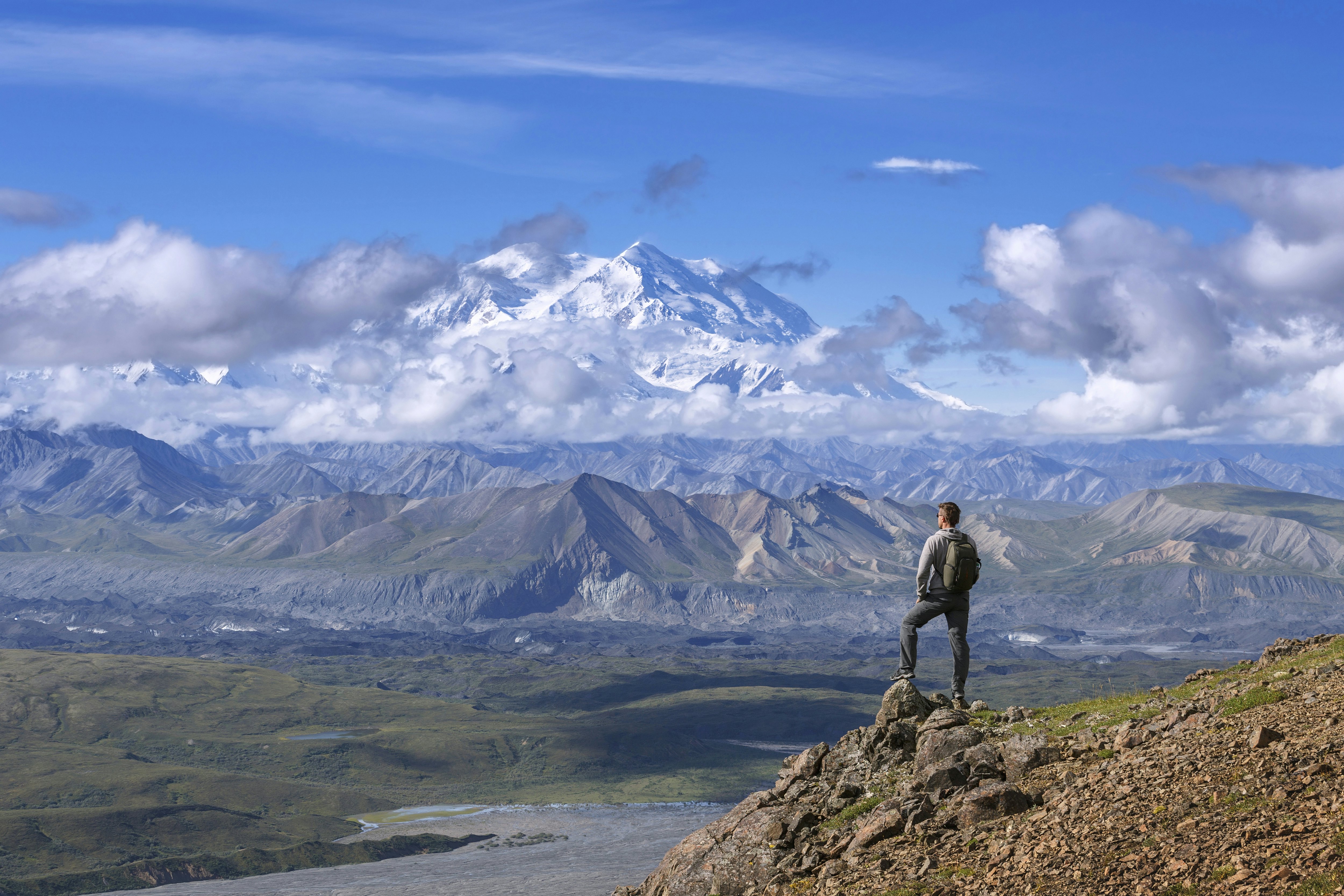 A hiker in shorts and a backpack stands atop a peak in Denali national park, Alaska