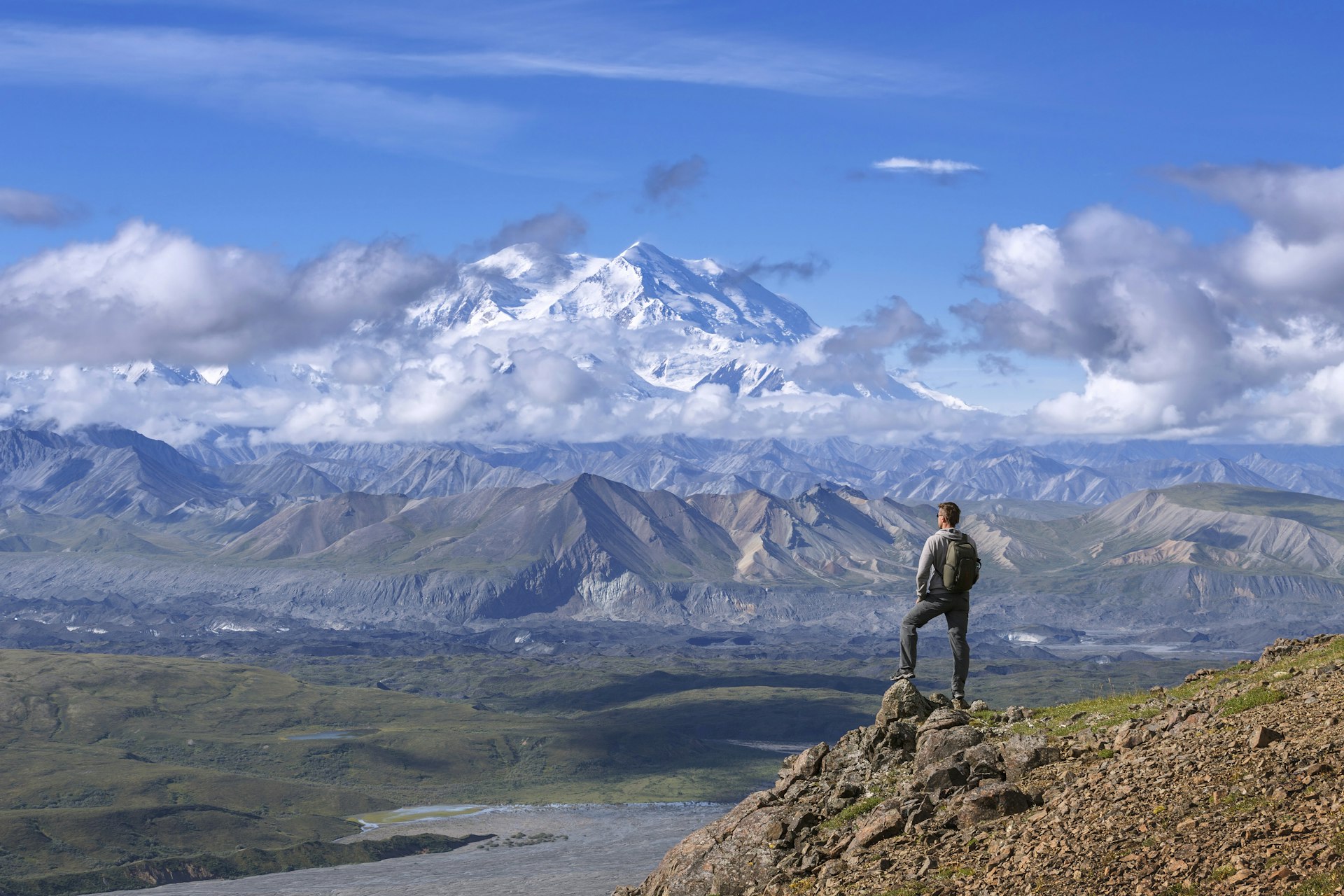 A hiker in shorts and a backpack stands atop a peak in Denali national park, Alaska