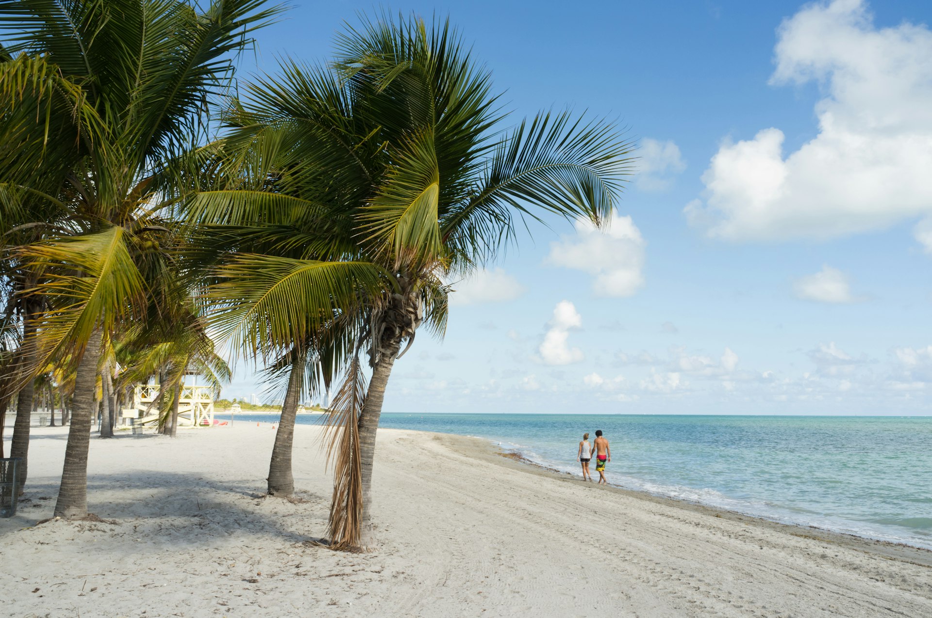 A couple in the distance, walking along a white-sand beach with palm trees 