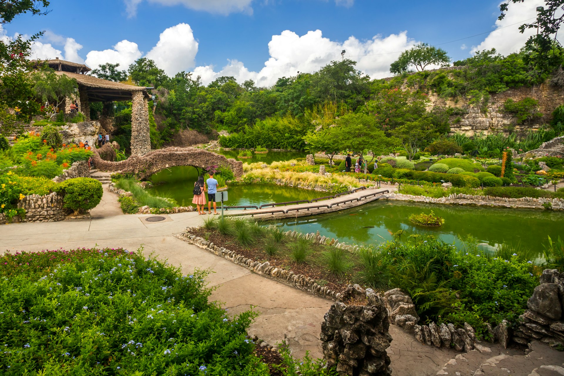 Two people stand on the edge of a series of ponds in a tranquil garden full of greenery