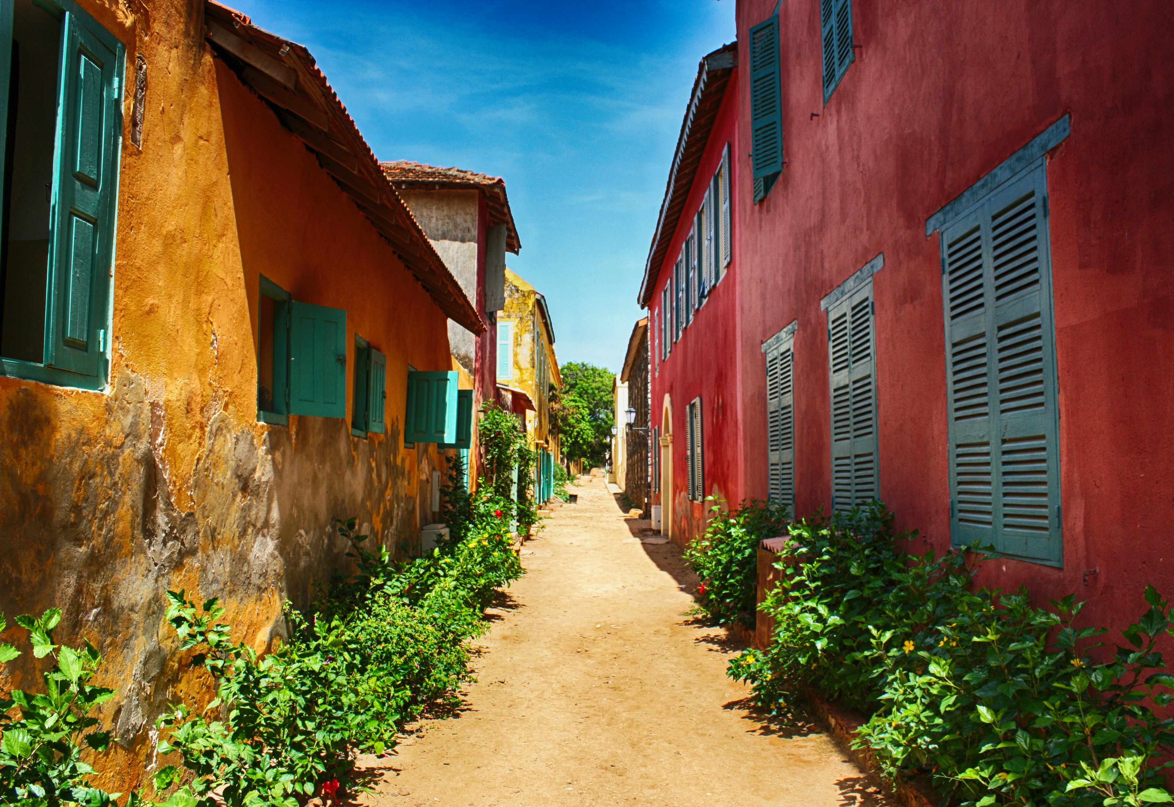 This image is looking straight down a sandy lane on Île de Gorée, with a bright red two-story building on the right and a yellow single-storey building on the left - both buildings have colourful shutters. Vegetation grows along the base of the buildlings