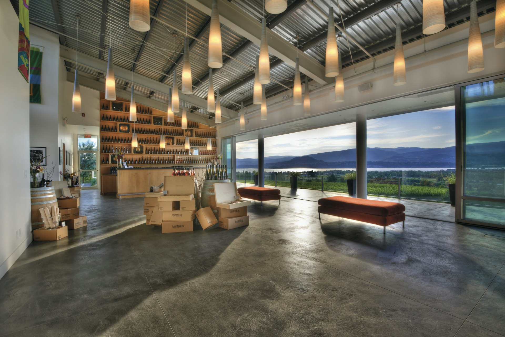 A large room with a bar at one end with shelves full of wine bottles. The floor-to-ceiling windows look out over vineyards